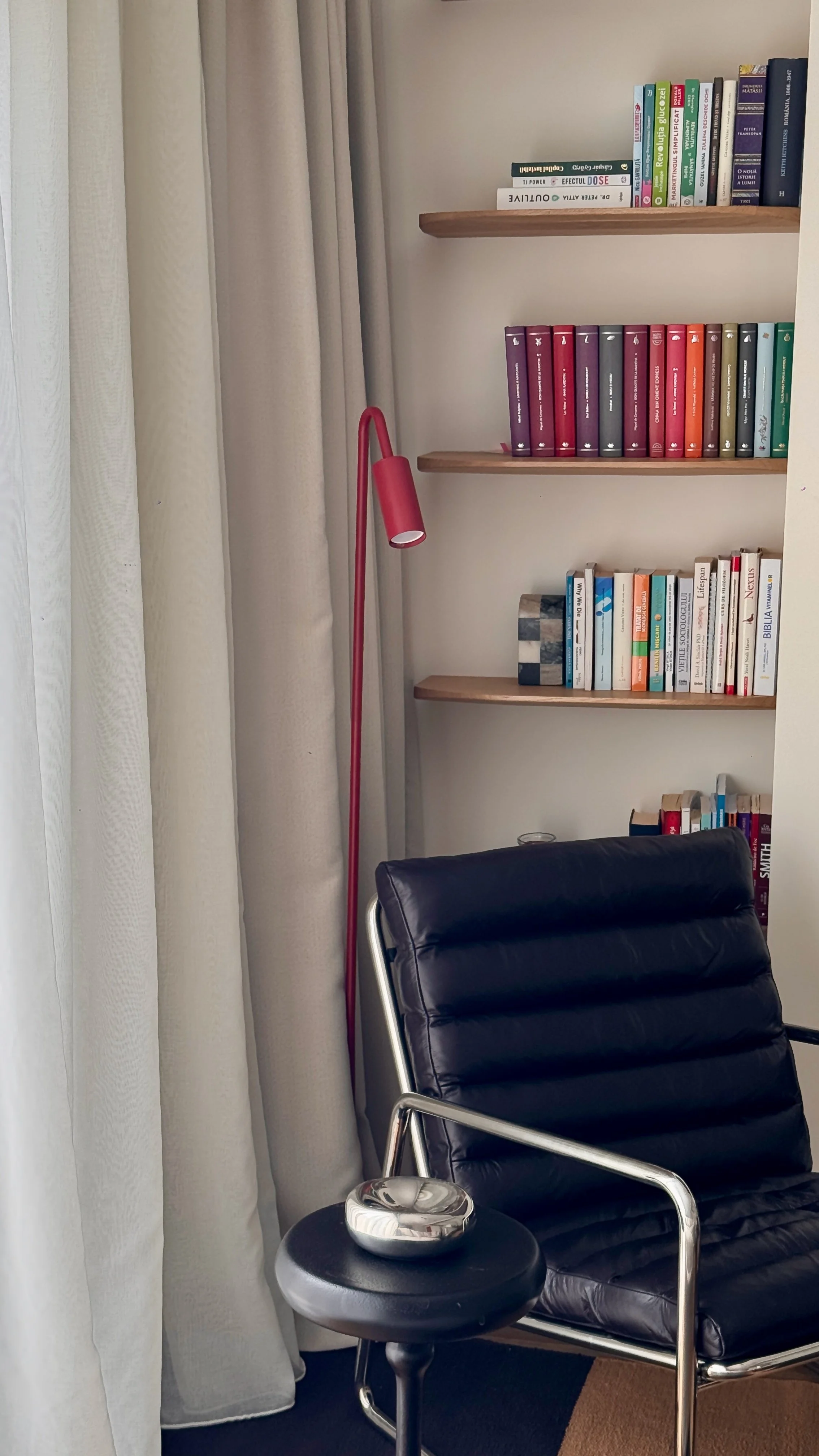 A black leather office chair with metal armrests and a small black side table with a glass ashtray on top, positioned next to a window with cream-colored curtains. Behind the chair are three wooden shelves filled with various books. A pink floor lamp