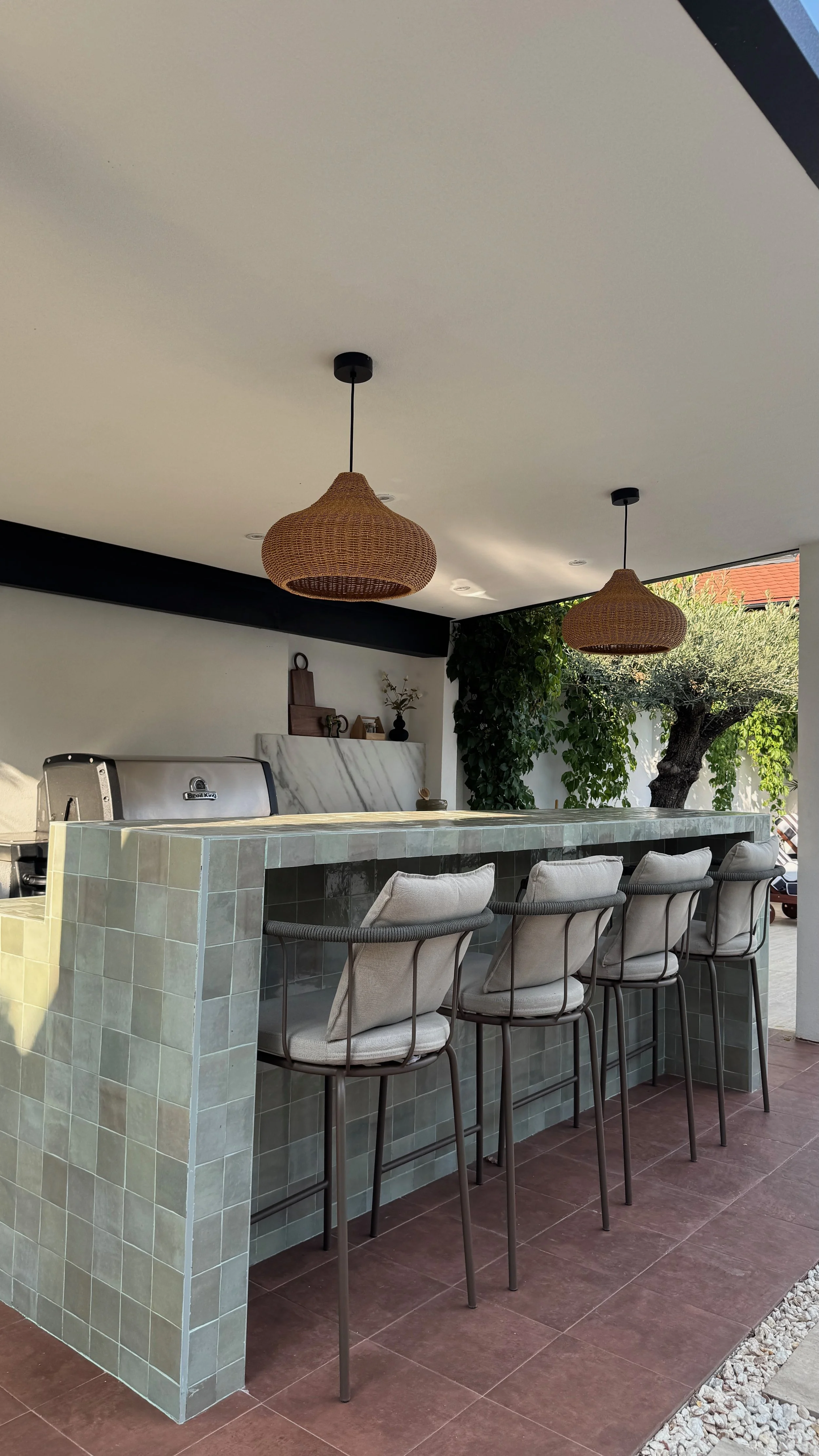 Outdoor bar with tiled counter and beige cushioned bar stools, pendant woven rattan light fixtures, potted plants, and trees in the background.