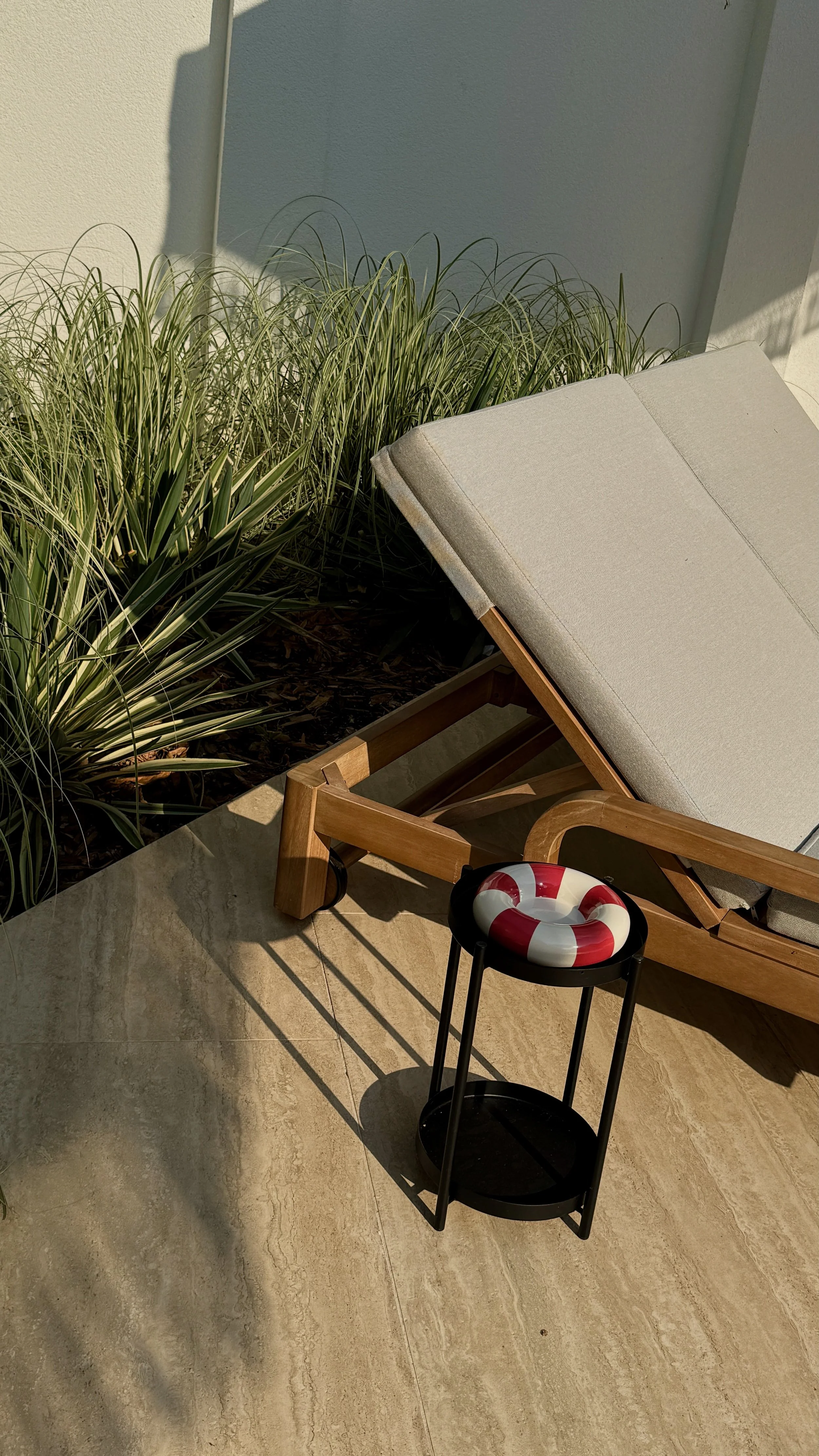 Outdoor patio scene with a beige cushioned lounge chair, a small black table with a decorative lifebuoy, green plants, and a wall with bright sunlight and shadows.
