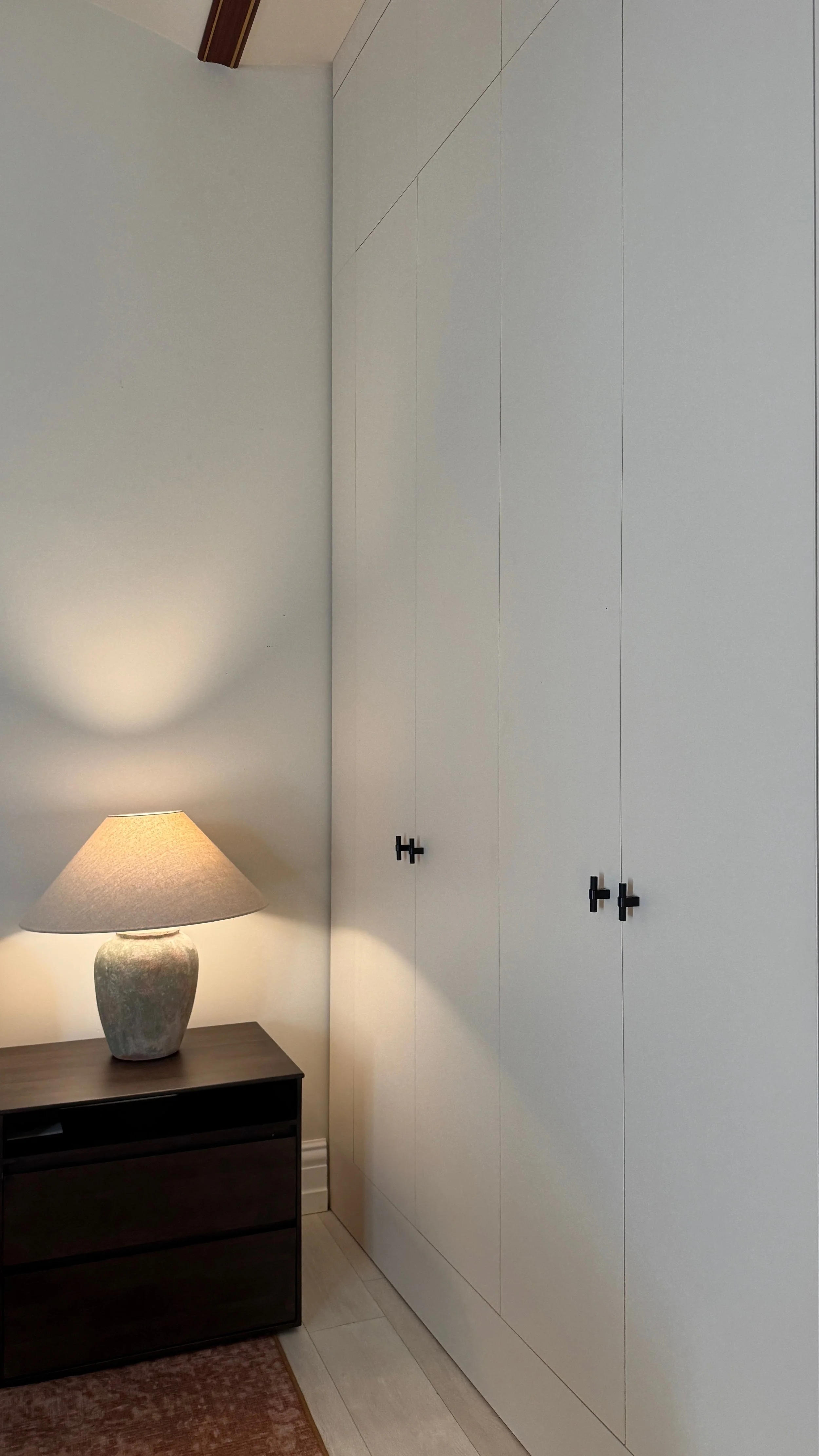 The corner of a bedroom with a mounted white closet, a small wooden nightstand, a table lamp, a patterned rug, and a beige tiled floor.