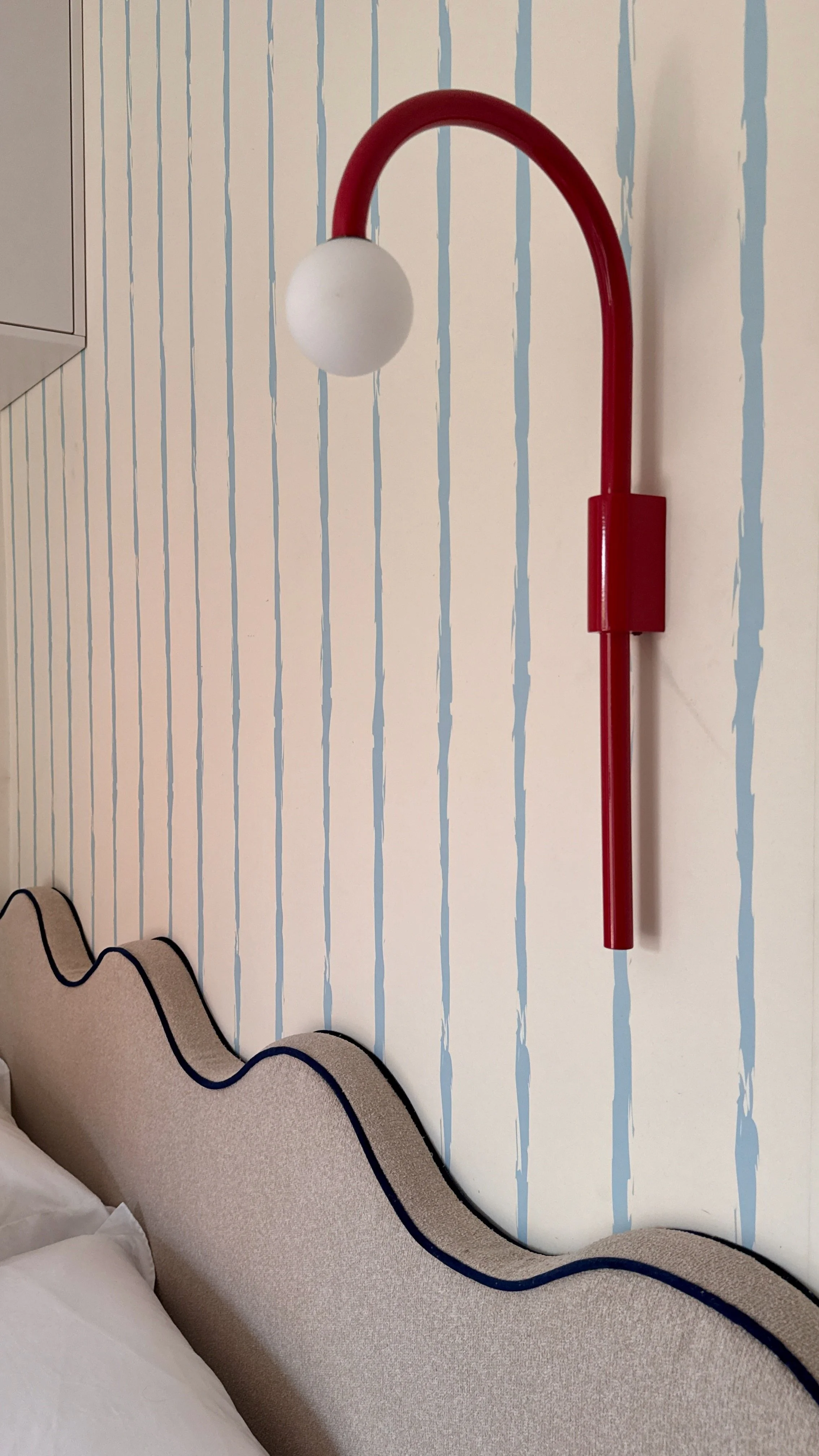 Red wall-mounted lamp with a white spherical bulb, attached to a blue and white striped wall in a bedroom.