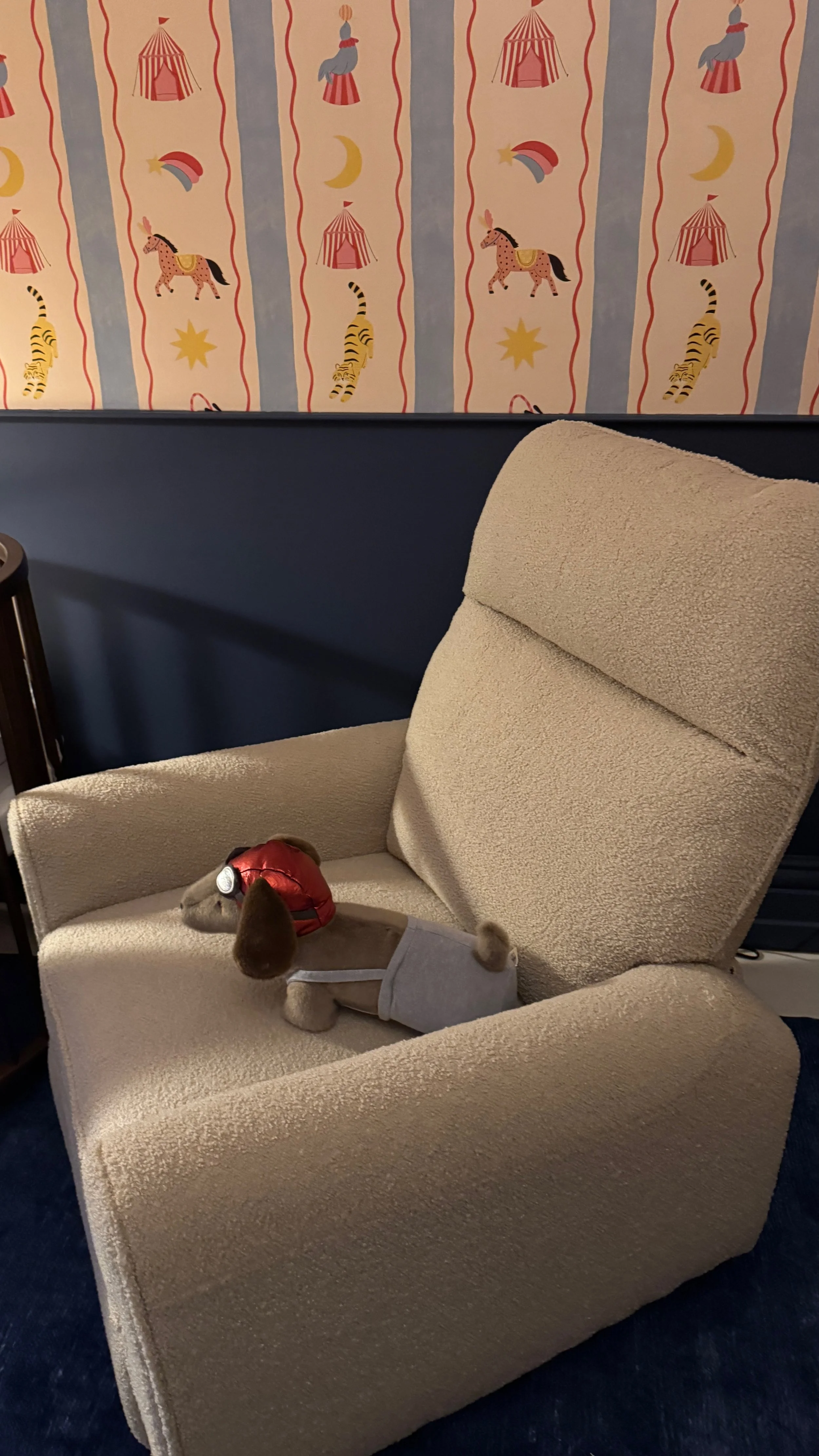 A plush dog wearing a gray outfit and red vest lies on a beige textured armchair in a room with a circus-themed wallpaper featuring tents, stars, moons, and carousel horses.