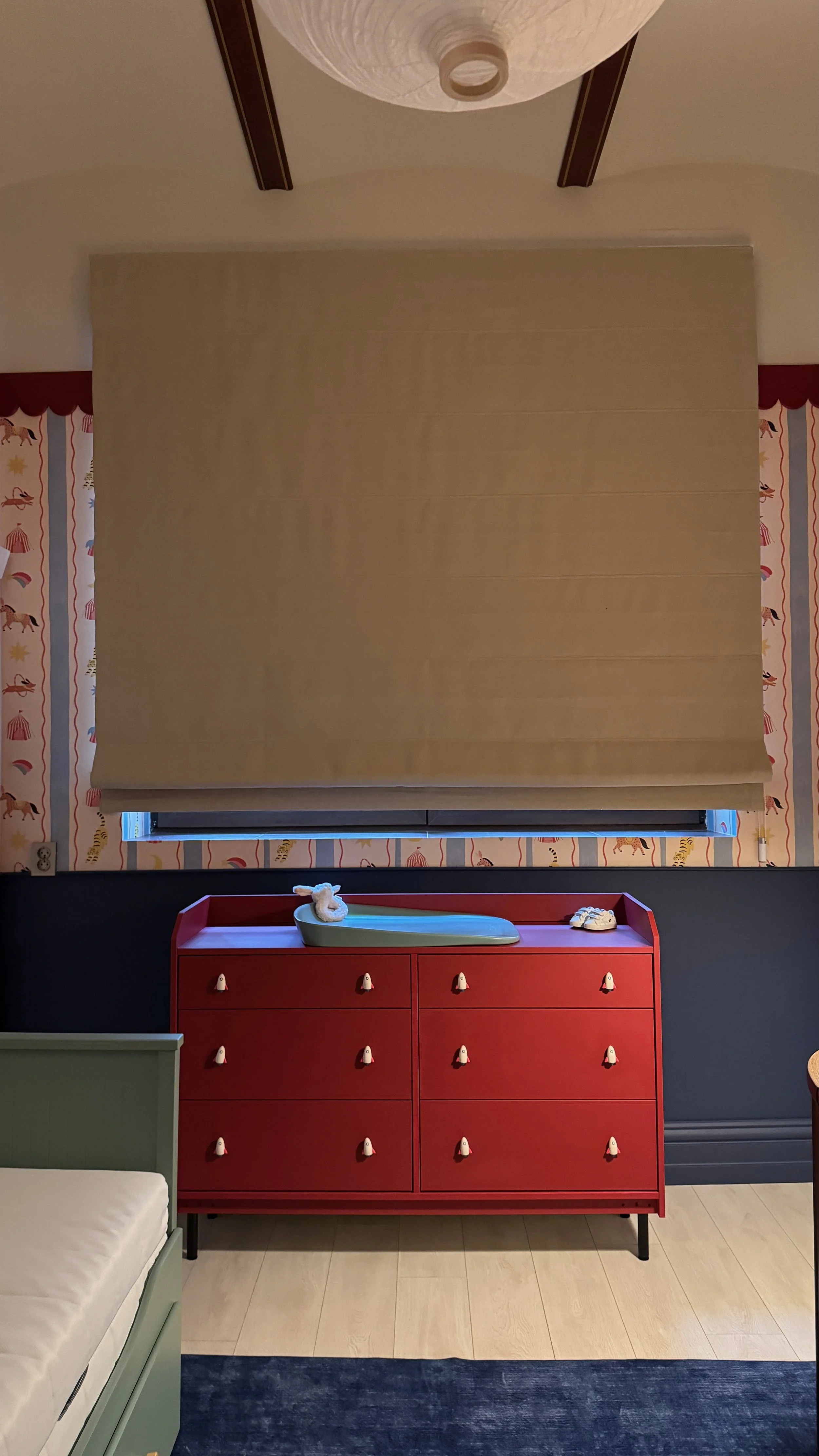 A red dresser with multiple drawers and white knobs, with a changing pad on top, in front of a window covered with beige shades. The wall behind features wallpaper with a red and white carnival or circus theme, including animals and tents.