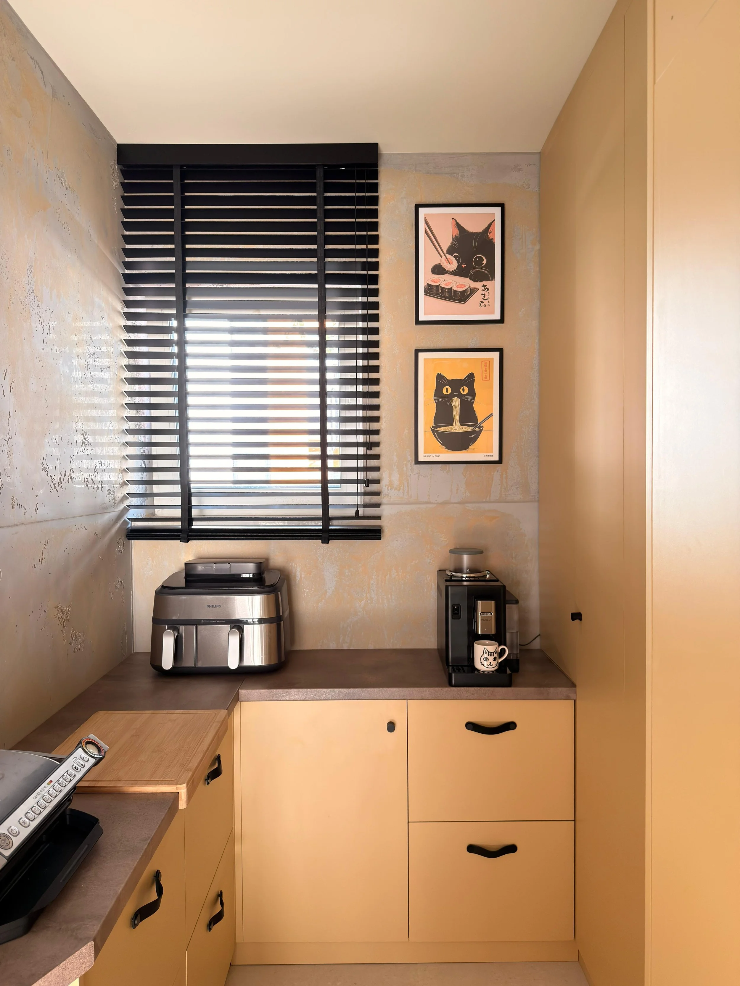 Black window blinds above a beige kitchen counter with kitchen appliances, framed cat-themed artwork on the wall, a coffee mug, and a cordless phone on the counter.