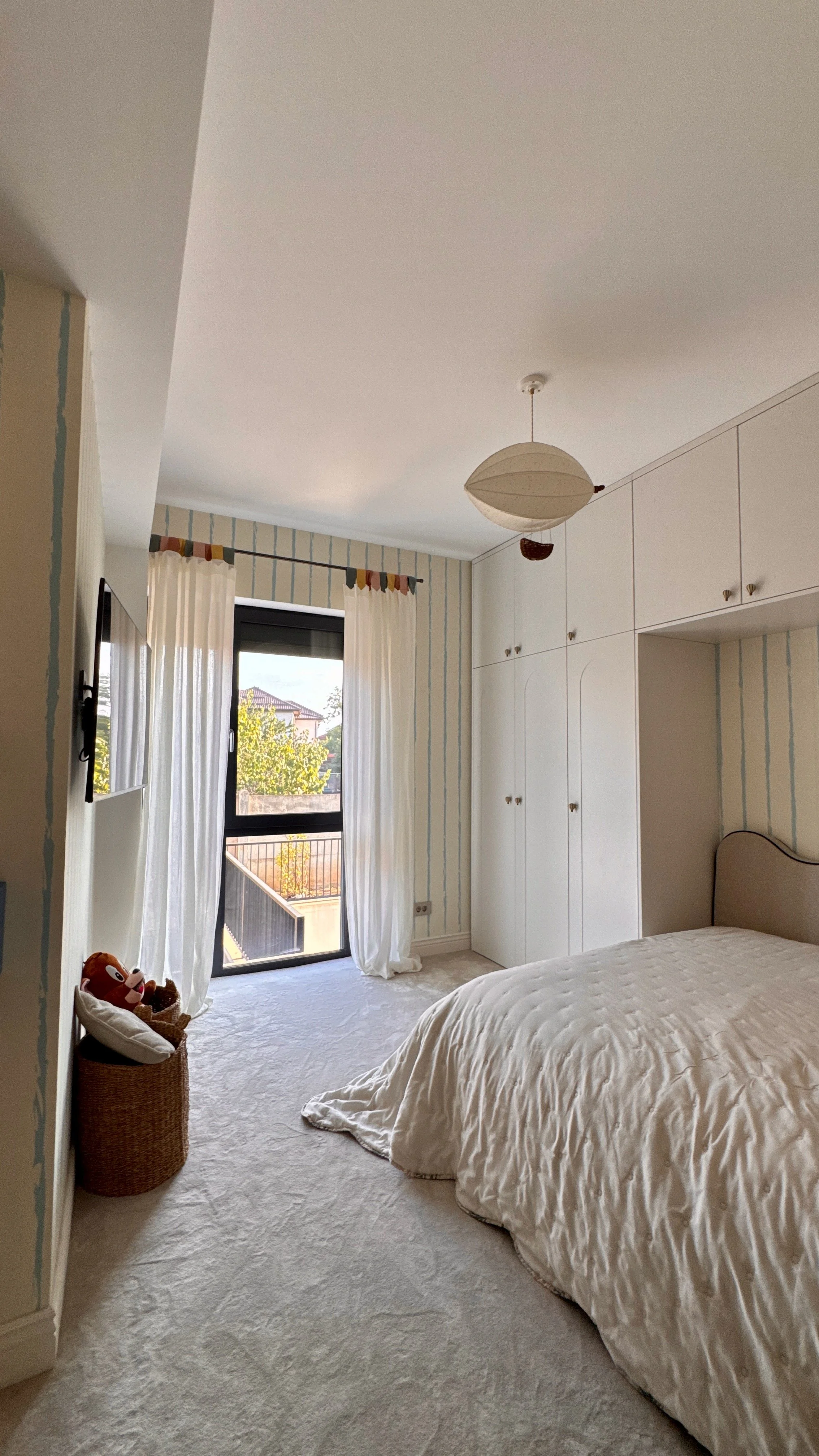 A bedroom with a large bed covered with a white duvet, tall white built-in closets, a window with white curtains, and a hanging ceiling lamp.