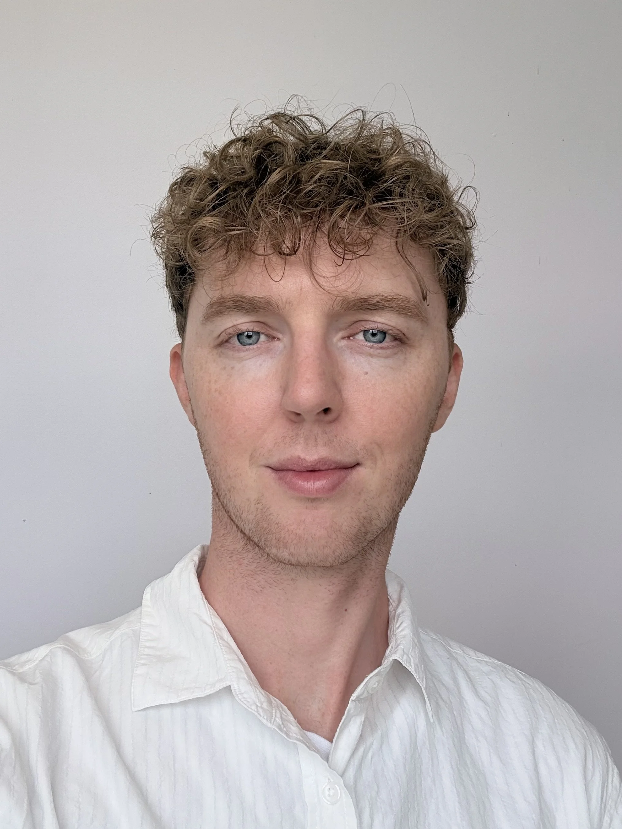 Portrait of a young man with light brown curly hair, wearing a white shirt, against a plain background.