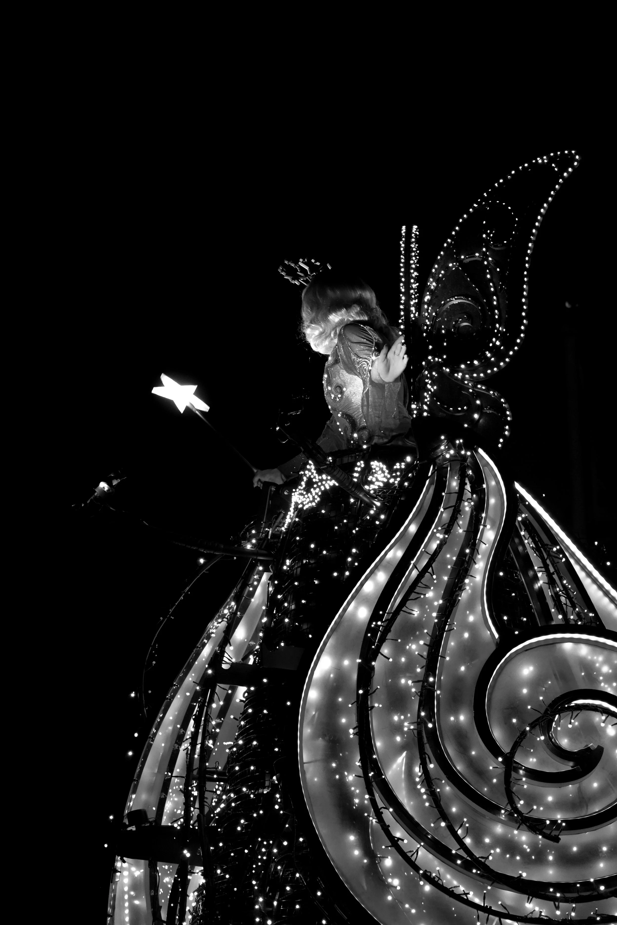 A child dressed as a fairy with illuminated wings riding in a decorated float with swirling patterns and lights at night.