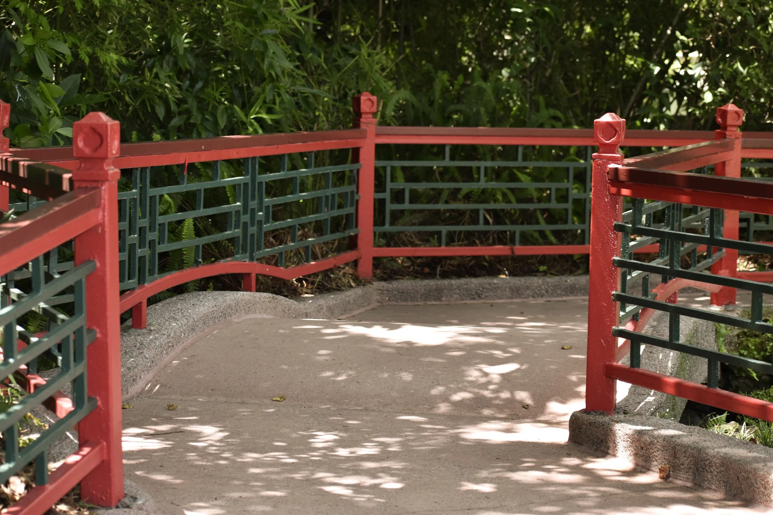 A small bridge with red and green railings over a pathway surrounded by green bushes and trees, with shadows cast on the path.