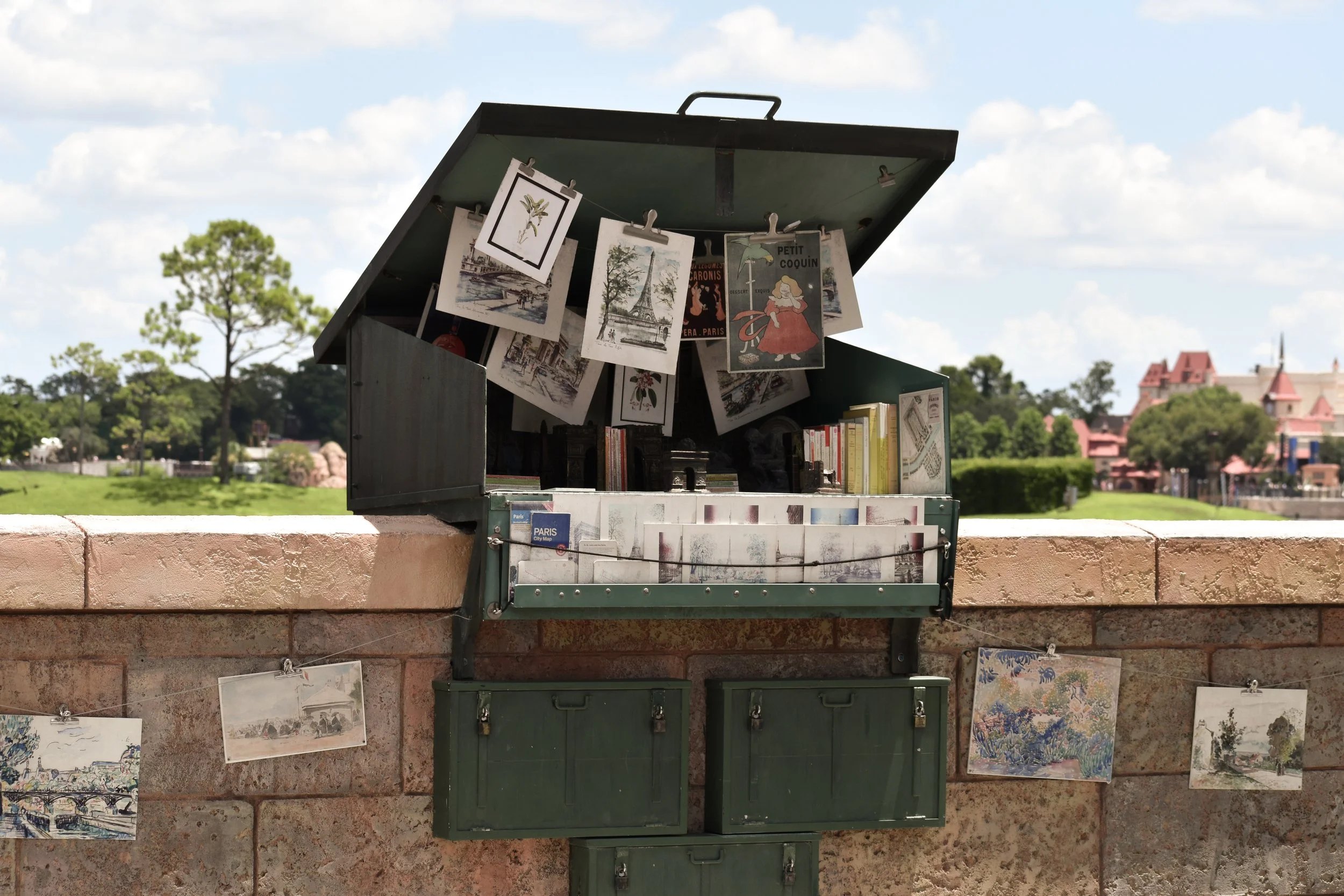 A small outdoor library on a brick wall with postcards and art prints displayed, overlooking a park with trees and buildings in the background.