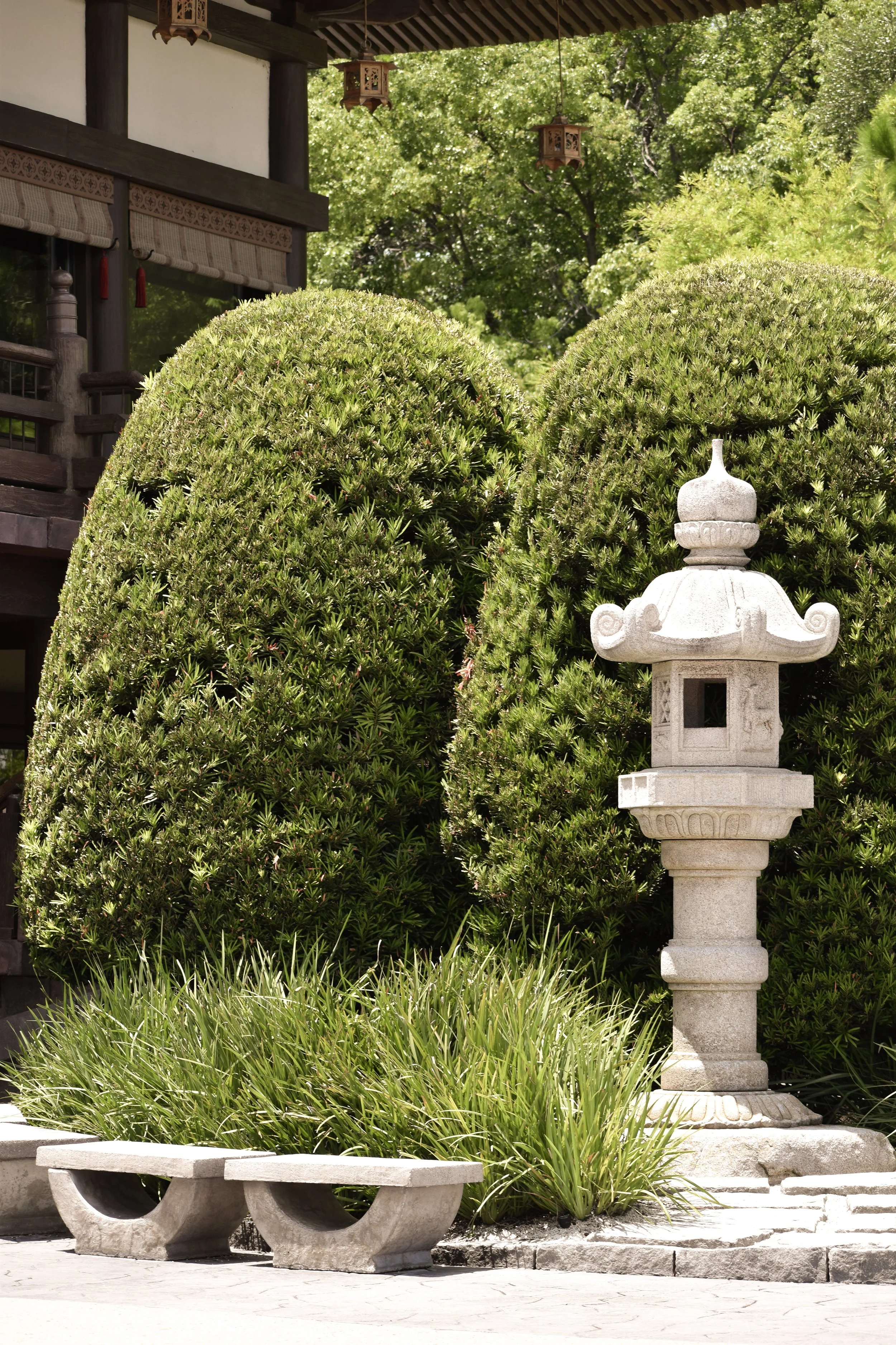 A stone Japanese lantern and a stone bench in front of neatly trimmed green bushes, with a traditional Japanese building and trees in the background.