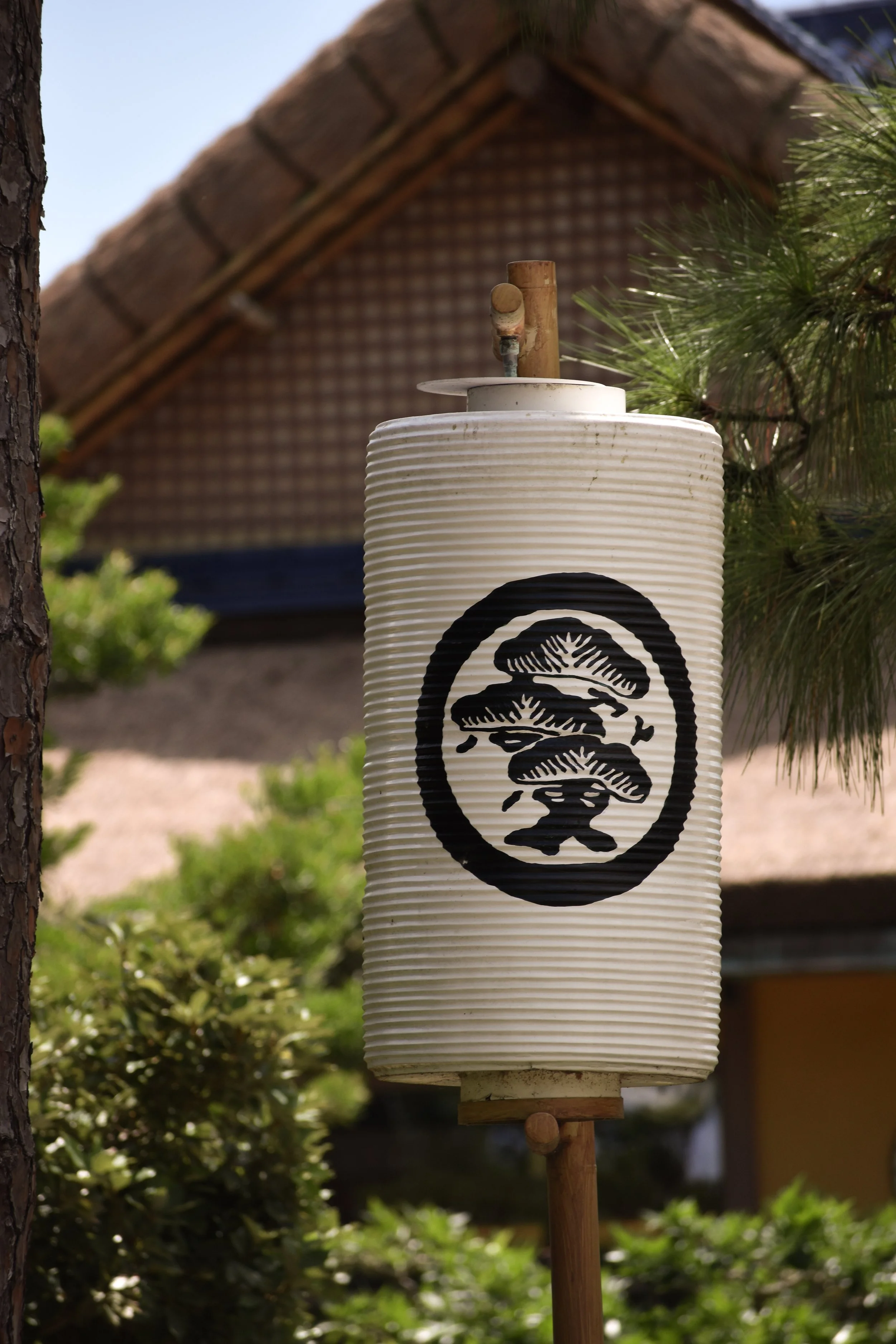 A traditional Japanese paper lantern with black design, hanging outdoors between trees and greenery.