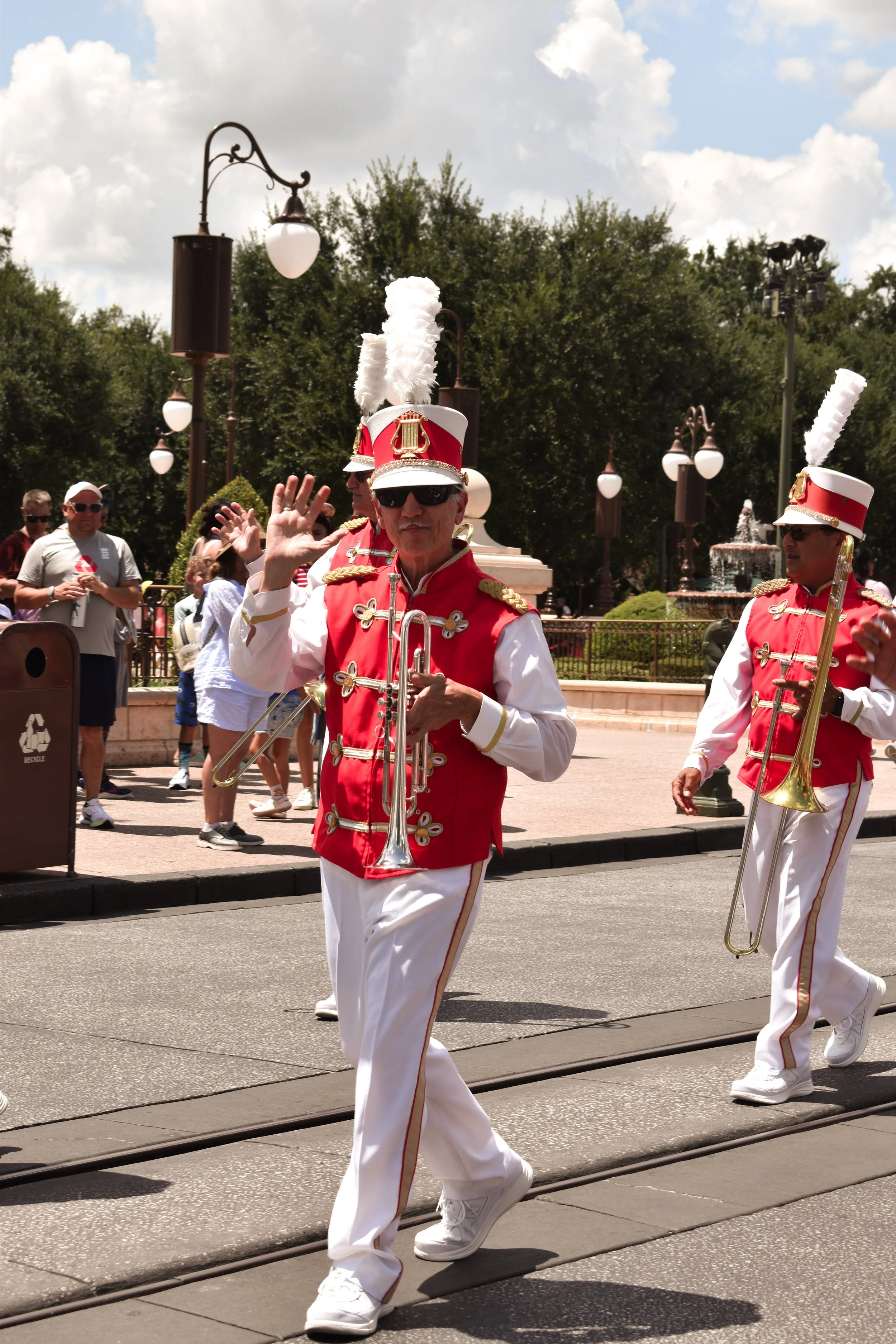 People dressed in marching band costumes walking on a parade route, with some spectators watching in the background, and a fountain and trees visible in a park setting.