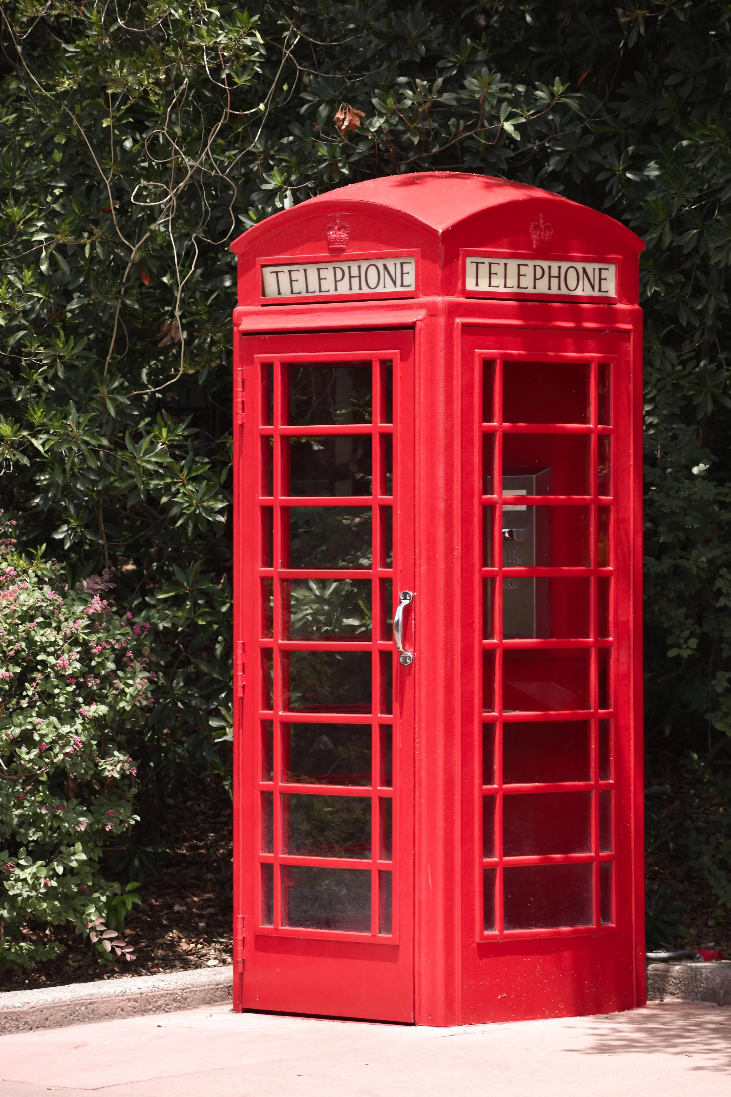 Red British-style telephone booth on sidewalk with greenery in background.