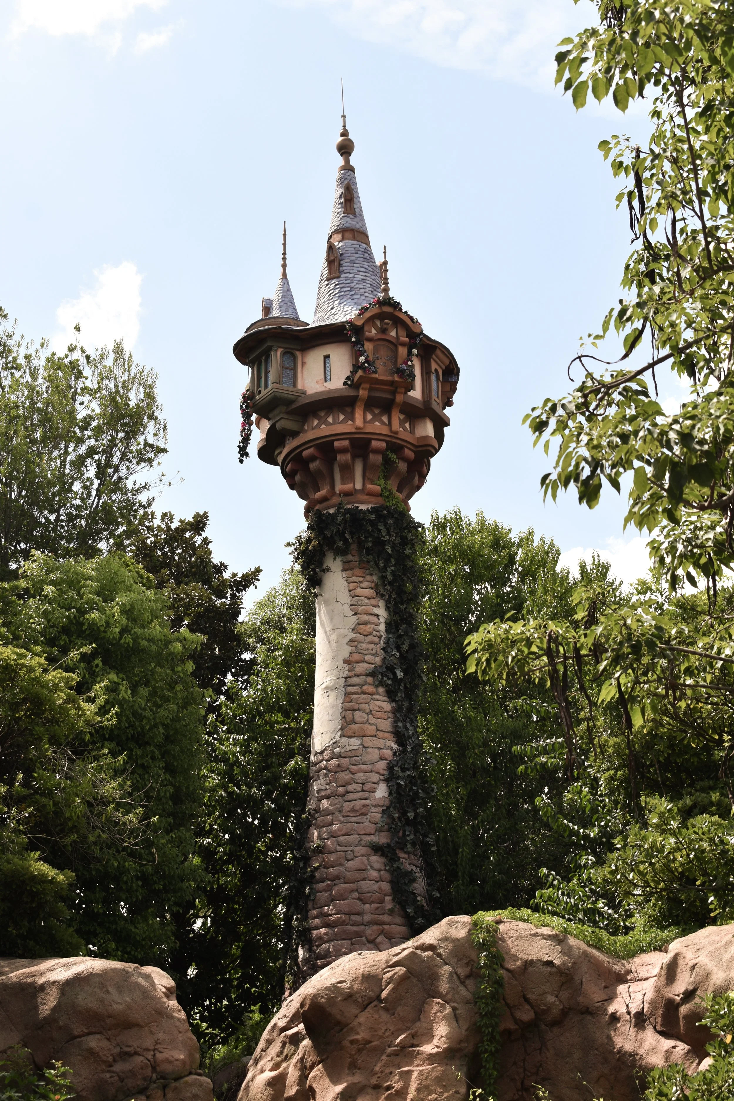 A tall stone tower with a whimsical, fairy-tale style topped with a castle-like structure, surrounded by trees and rocks.