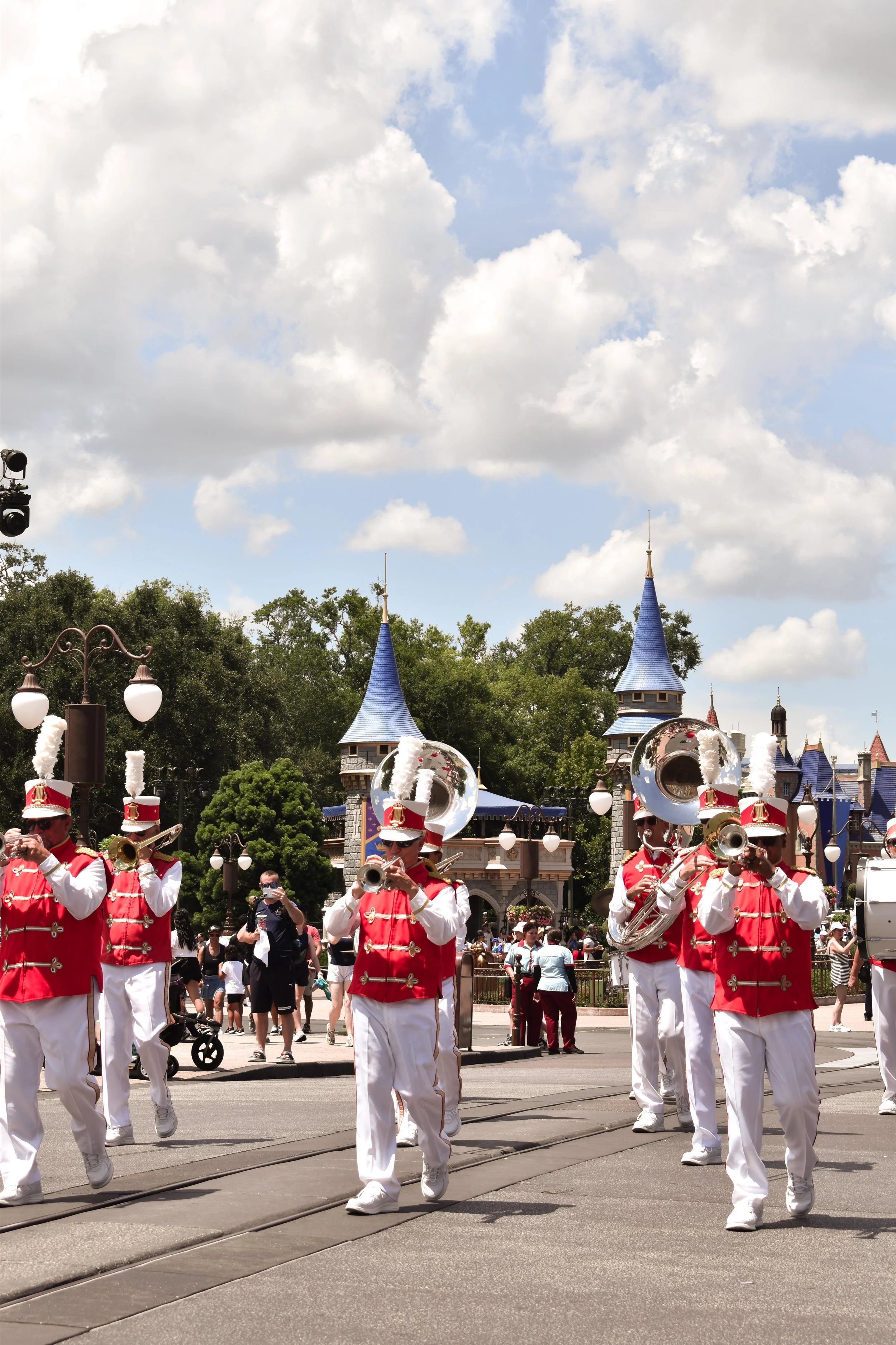 Marching band performers in red and white uniforms playing instruments during a parade at Disneyland with Sleeping Beauty Castle in the background.
