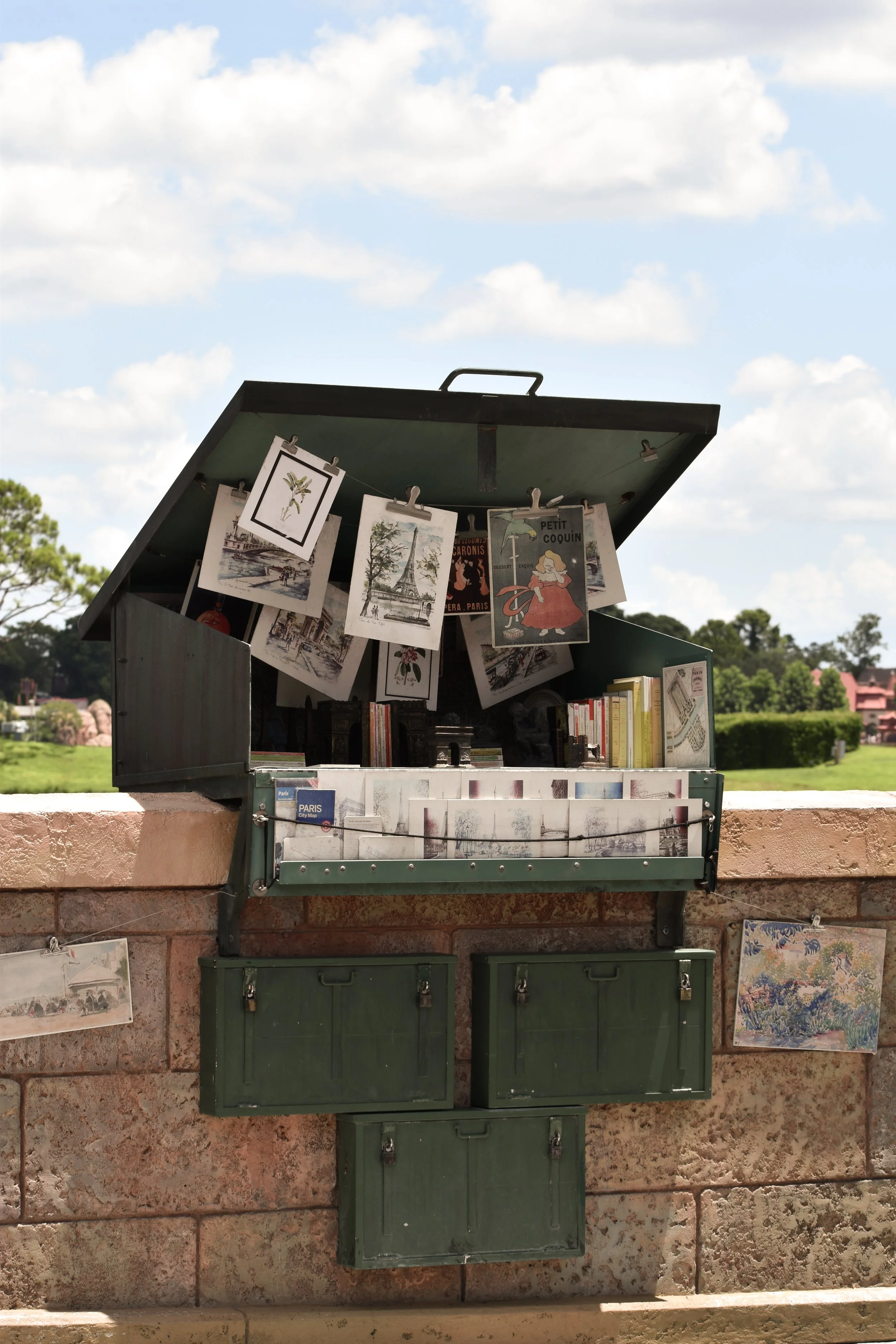 A small, vintage green display case mounted on a brick wall, holding postcards and artwork, with more postcards and artwork hanging below, against a background of blue sky with clouds and green trees and fields.
