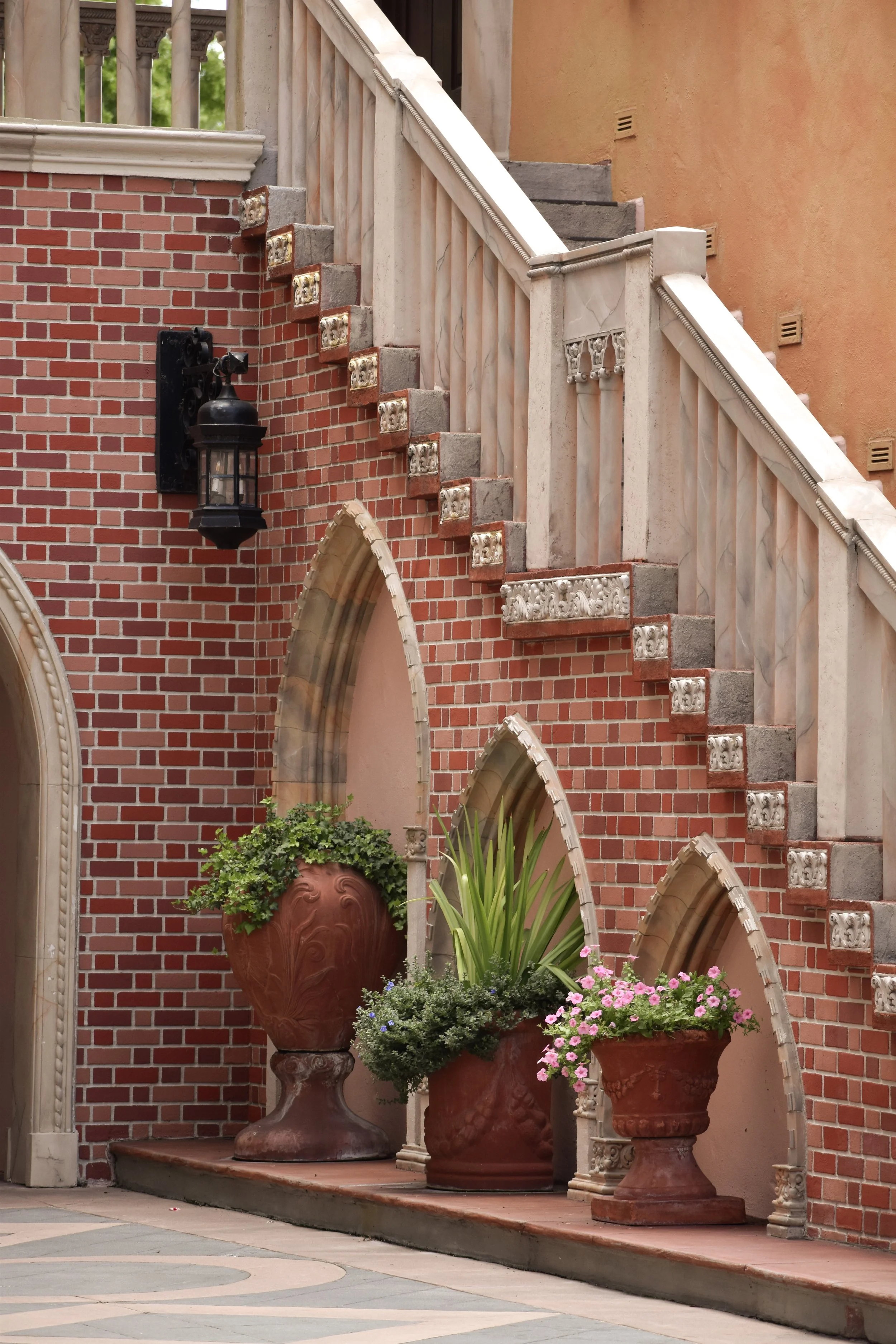 Potted plants and flowers arranged on a ledge in front of a brick building with an arched doorway and decorative staircase.