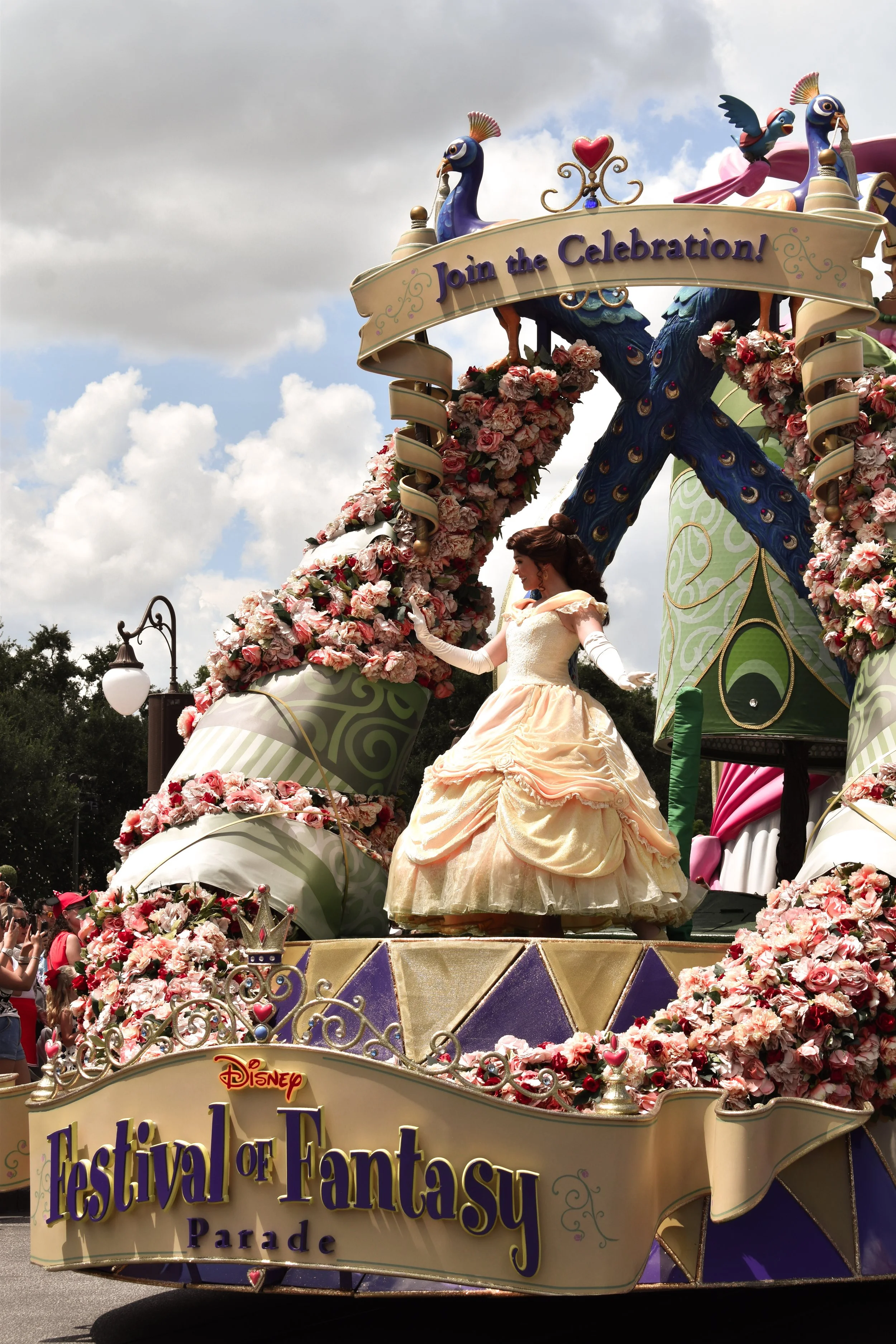 A parade float decorated with pink and white flowers, featuring a Cinderella doll in a peach-colored ball gown. The float has a sign that reads "Join the Celebration!" and another that says "Festival of Fantasy Parade."