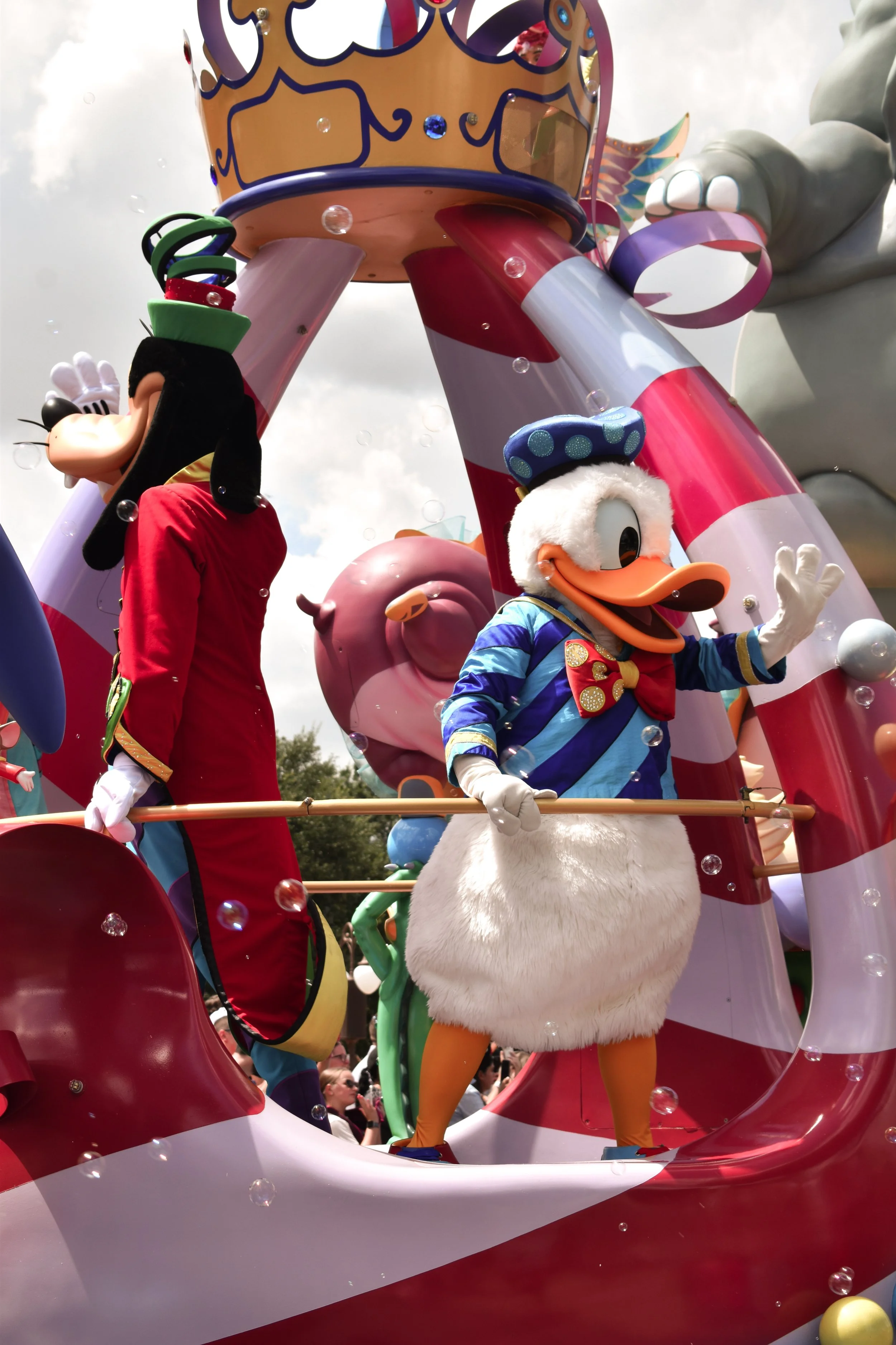 Costumed characters of Mickey Mouse and Donald Duck participating in a parade float, with a large pink ear and candy cane striped decorations in the background, and crowd visible at the bottom.