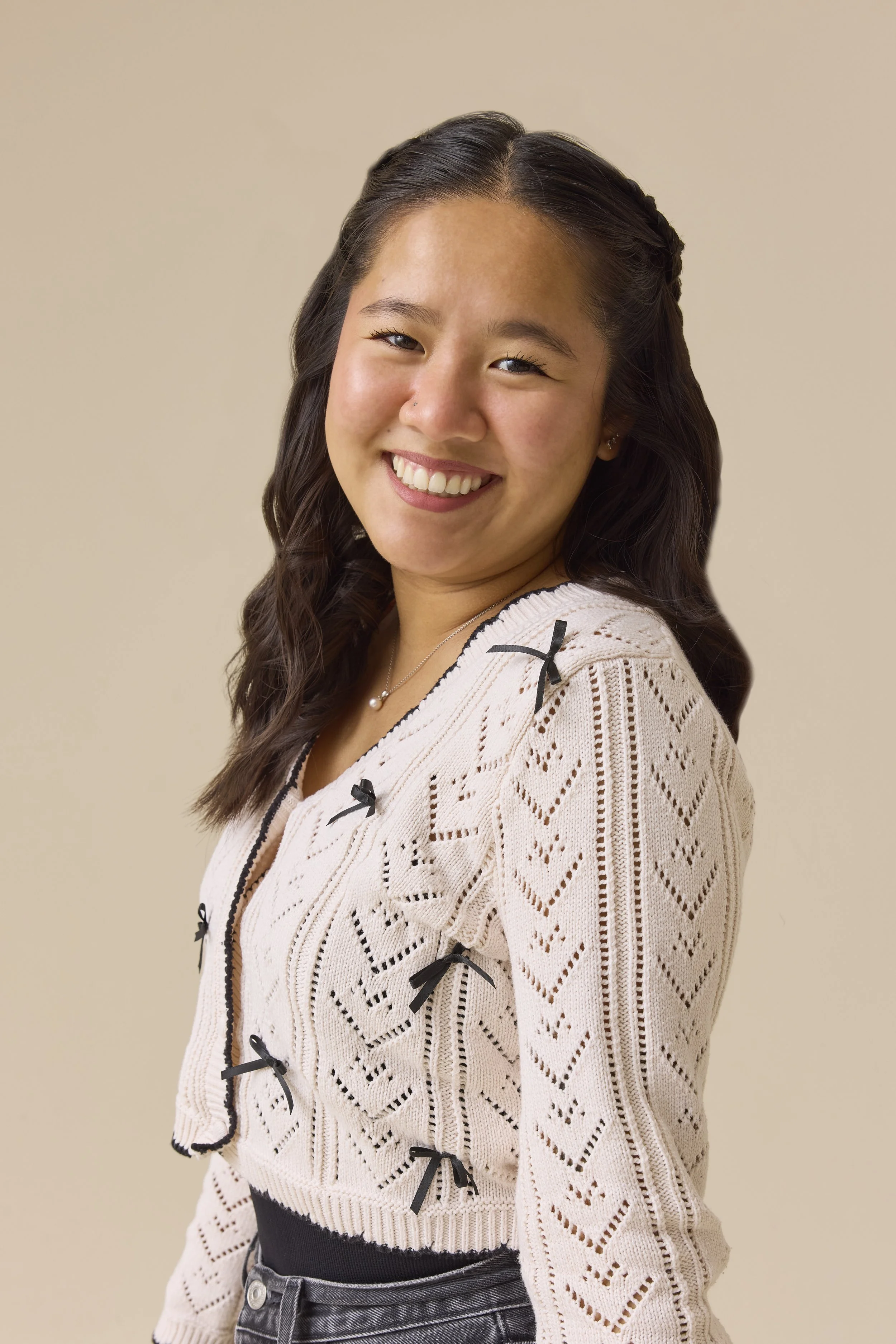 A young woman with dark wavy hair smiling at the camera, wearing a beige knit sweater with black bows and a black skirt, standing against a plain beige background.