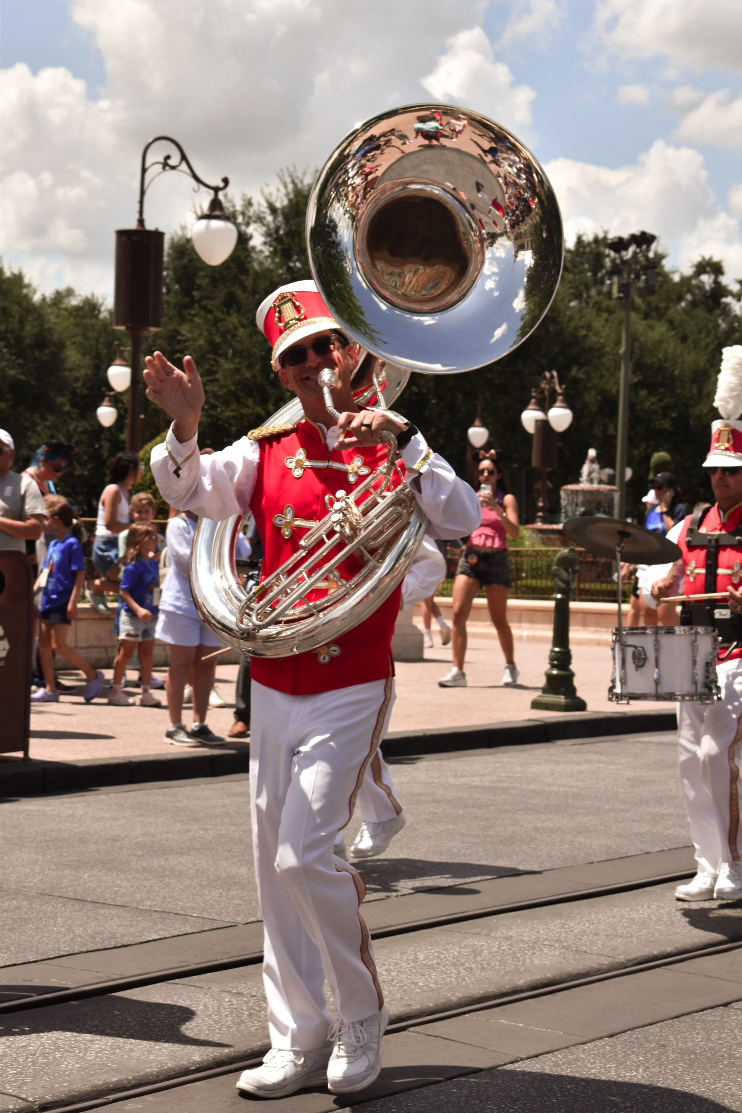 A marching band member playing a sousaphone during a parade, dressed in a red and white uniform with a red hat and gold accents, on a sunny day with onlookers in the background.