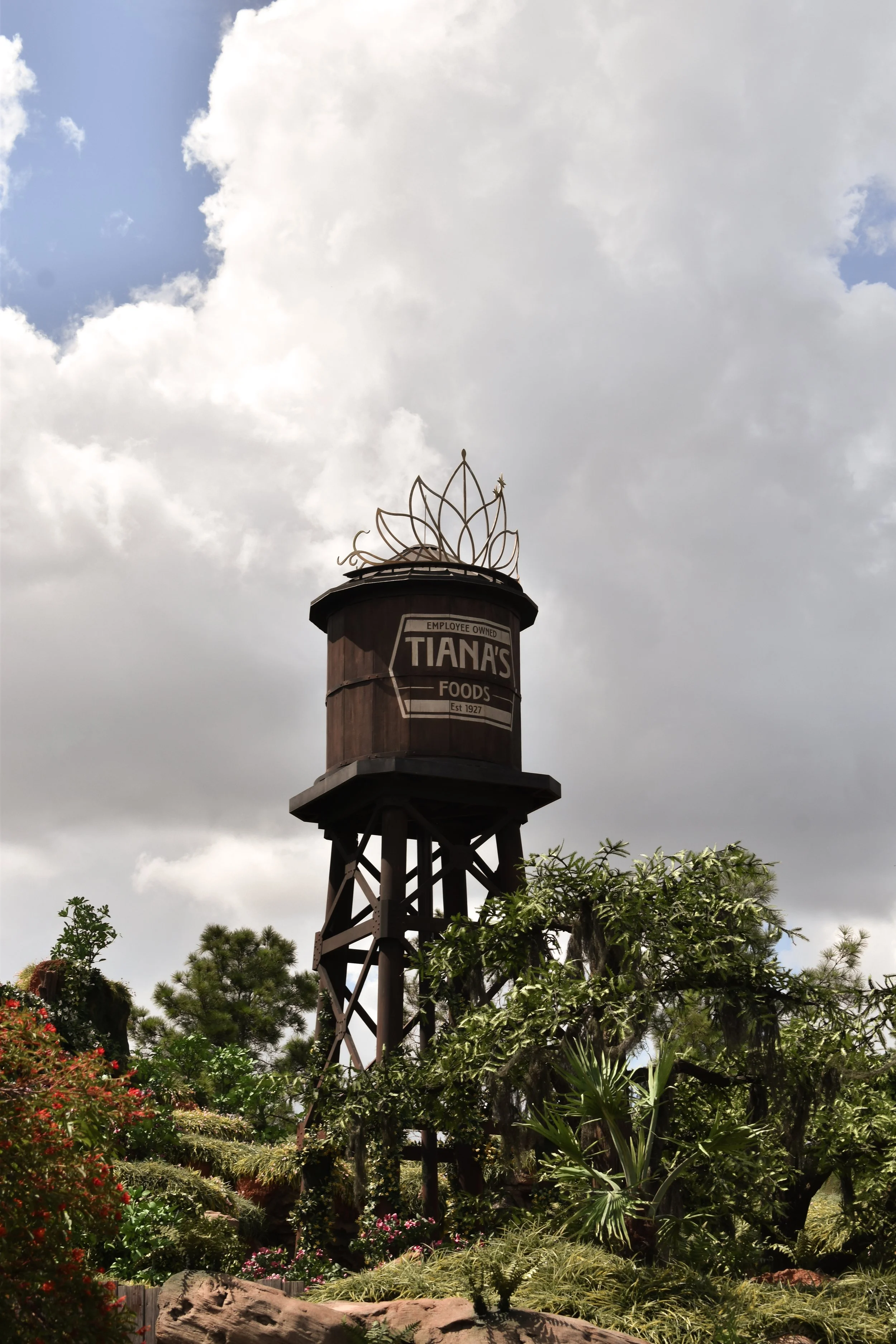 Wooden water tower with Tiana's Foods sign, topped with metal decorative flower, surrounded by lush greenery and cloudy sky