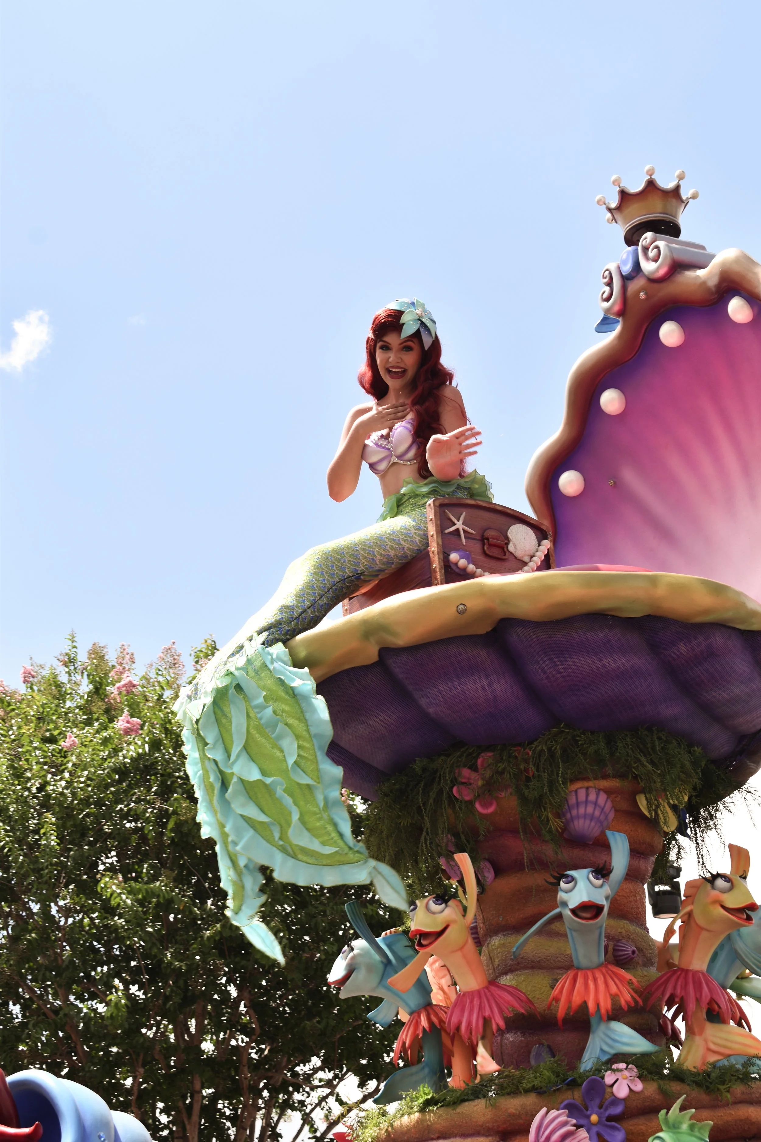 A parade float featuring a mermaid character with red hair, a purple shell top, and a green tail, sitting on a large decorative shell with sea creatures at the bottom, against a clear blue sky.