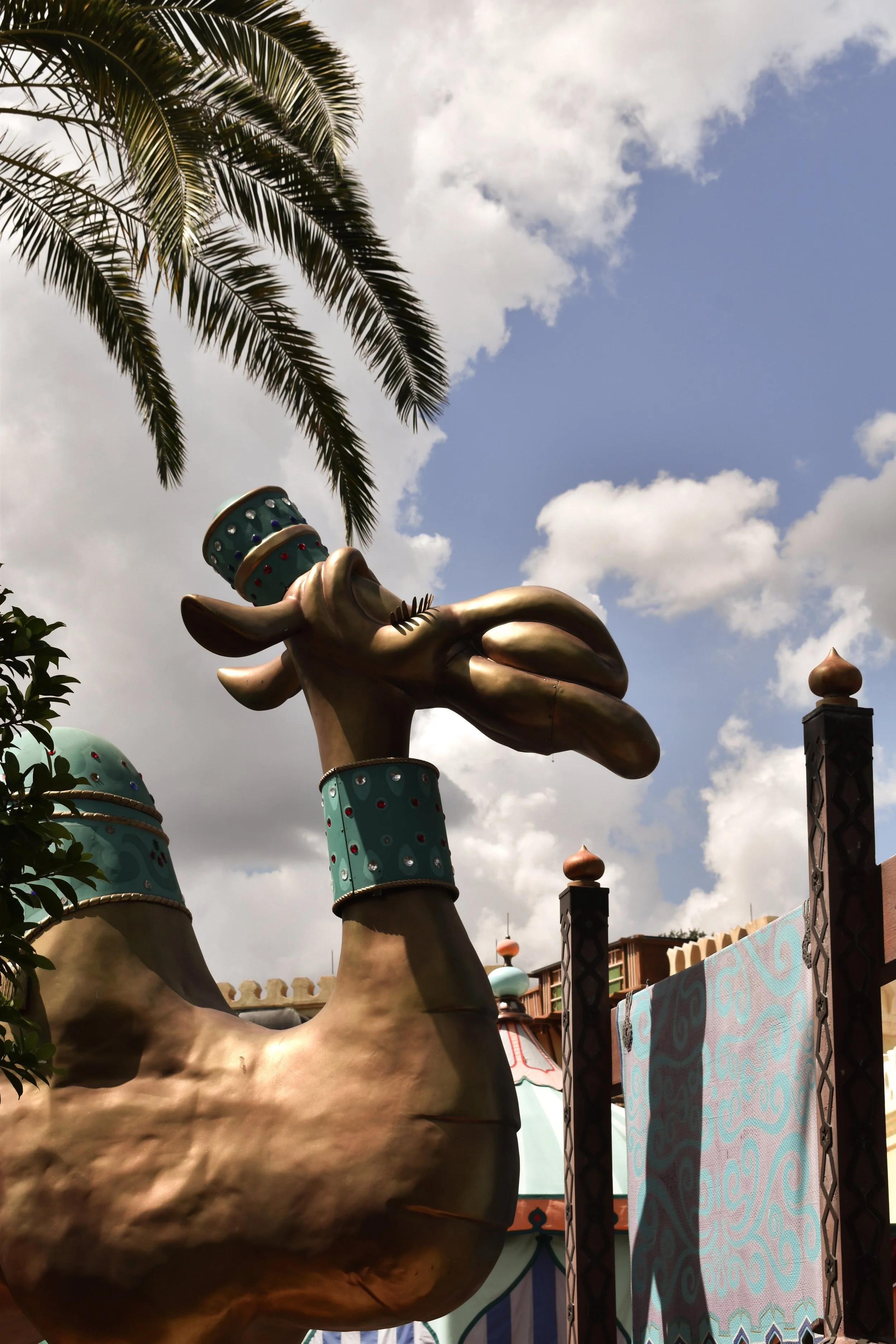 A large decorative sculpture of a camel with a crown and collar, with a palm tree and a partly cloudy sky in the background.