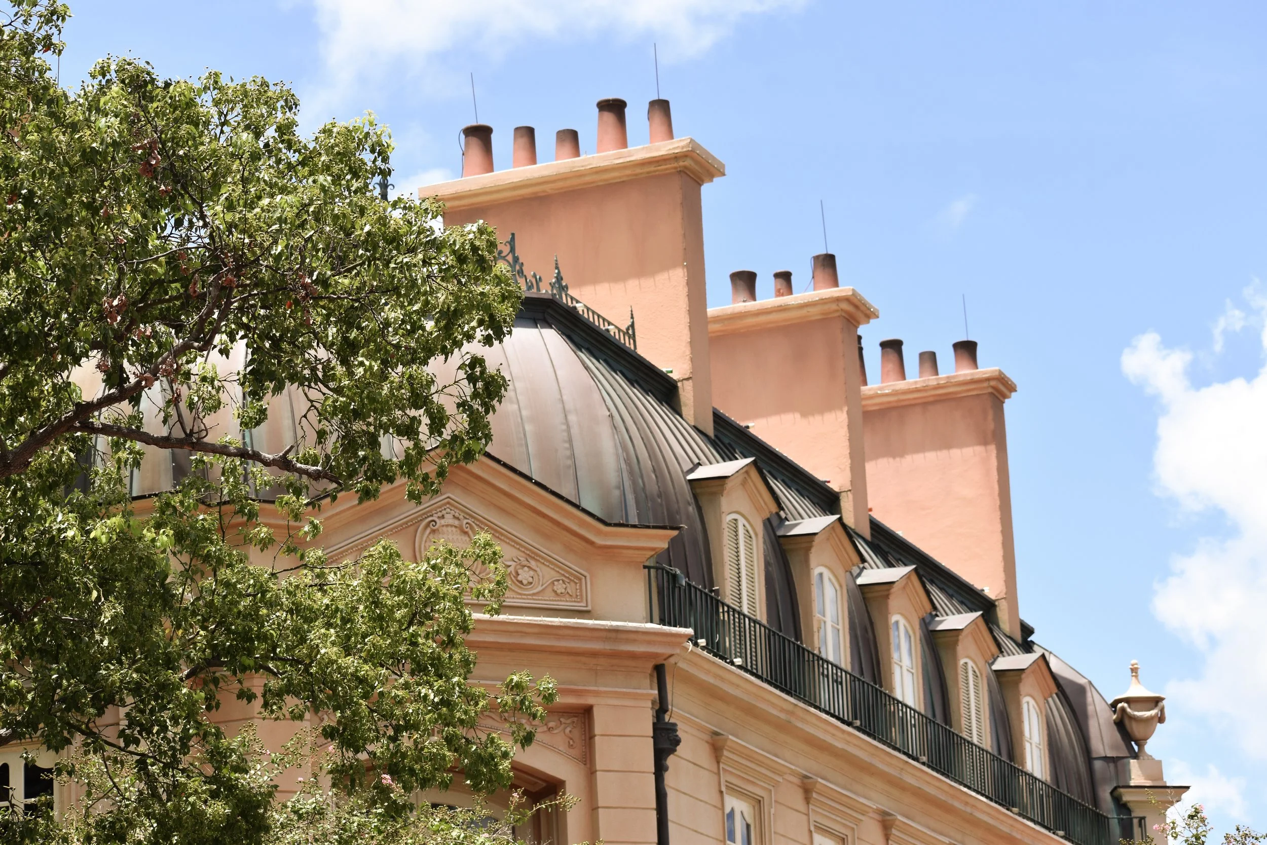 Top part of a historic building with multiple chimneys, pink walls, black metal roof, and a tree partially in front, set against a blue sky with some clouds.