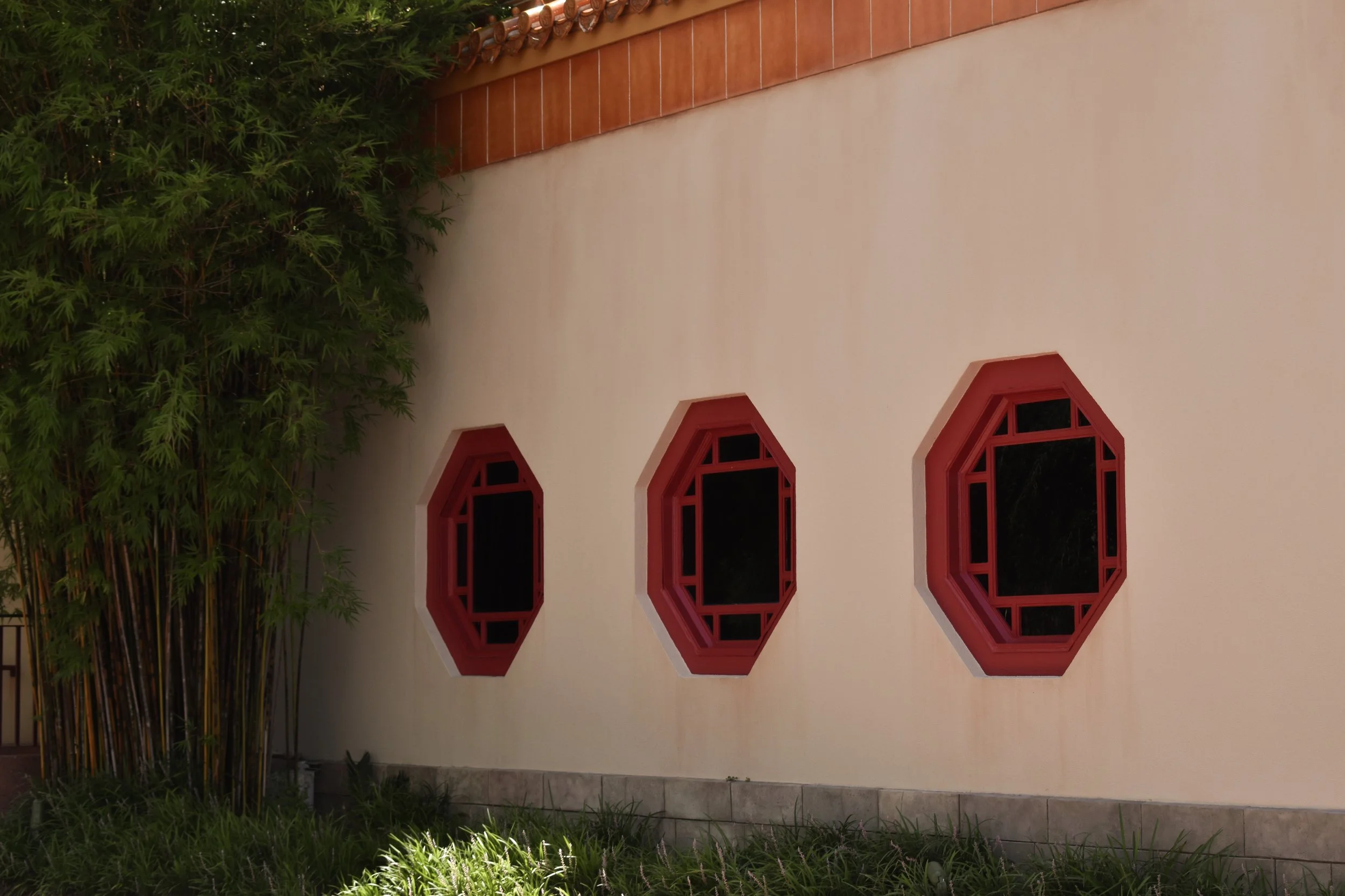 Exterior wall with three octagonal windows framed in red, next to green bamboo plants and grass.