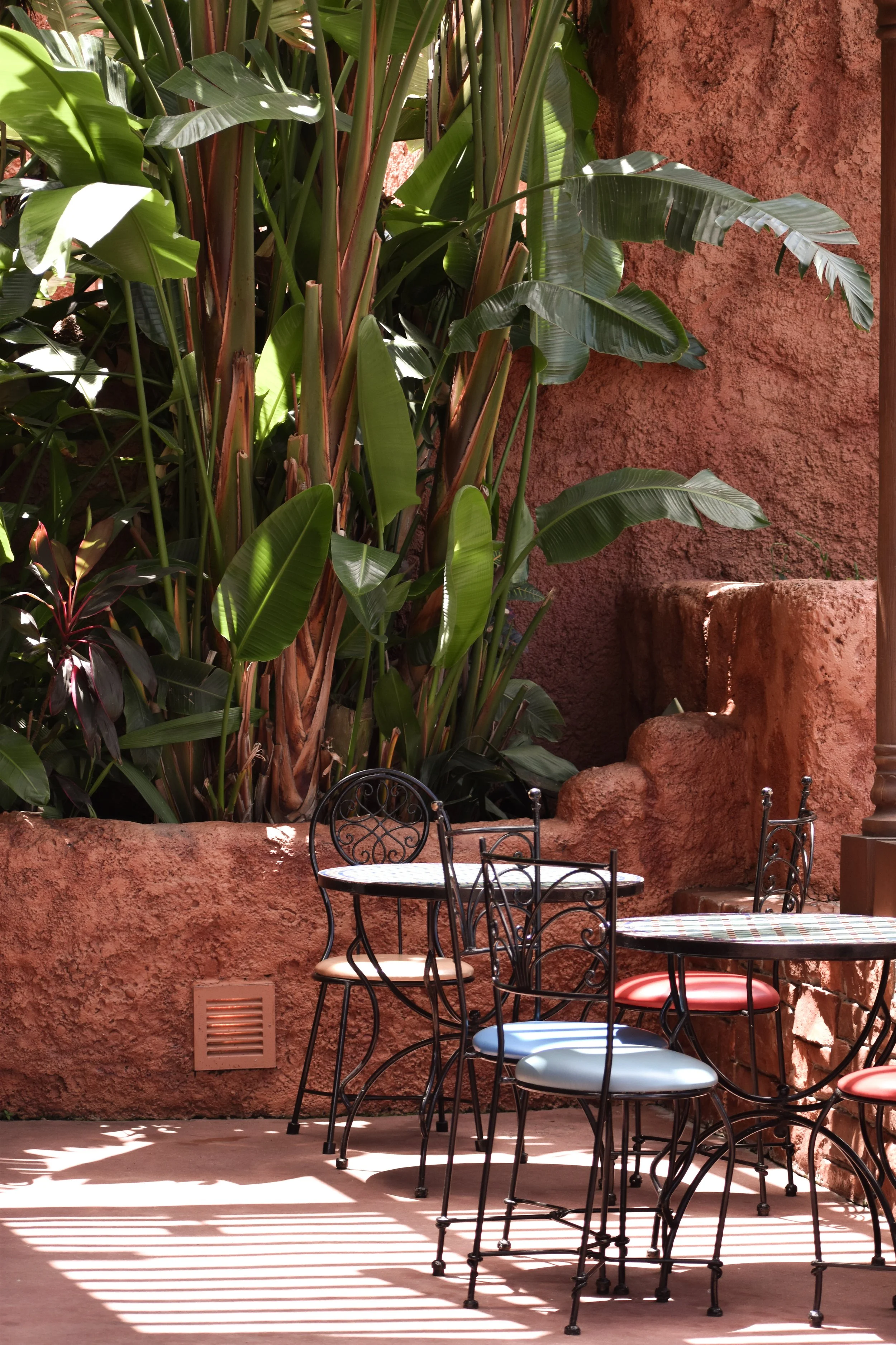 Outdoor patio area with iron chairs and tables near a terracotta-colored stucco wall, lush green tropical plants, and sunlight casting shadows.