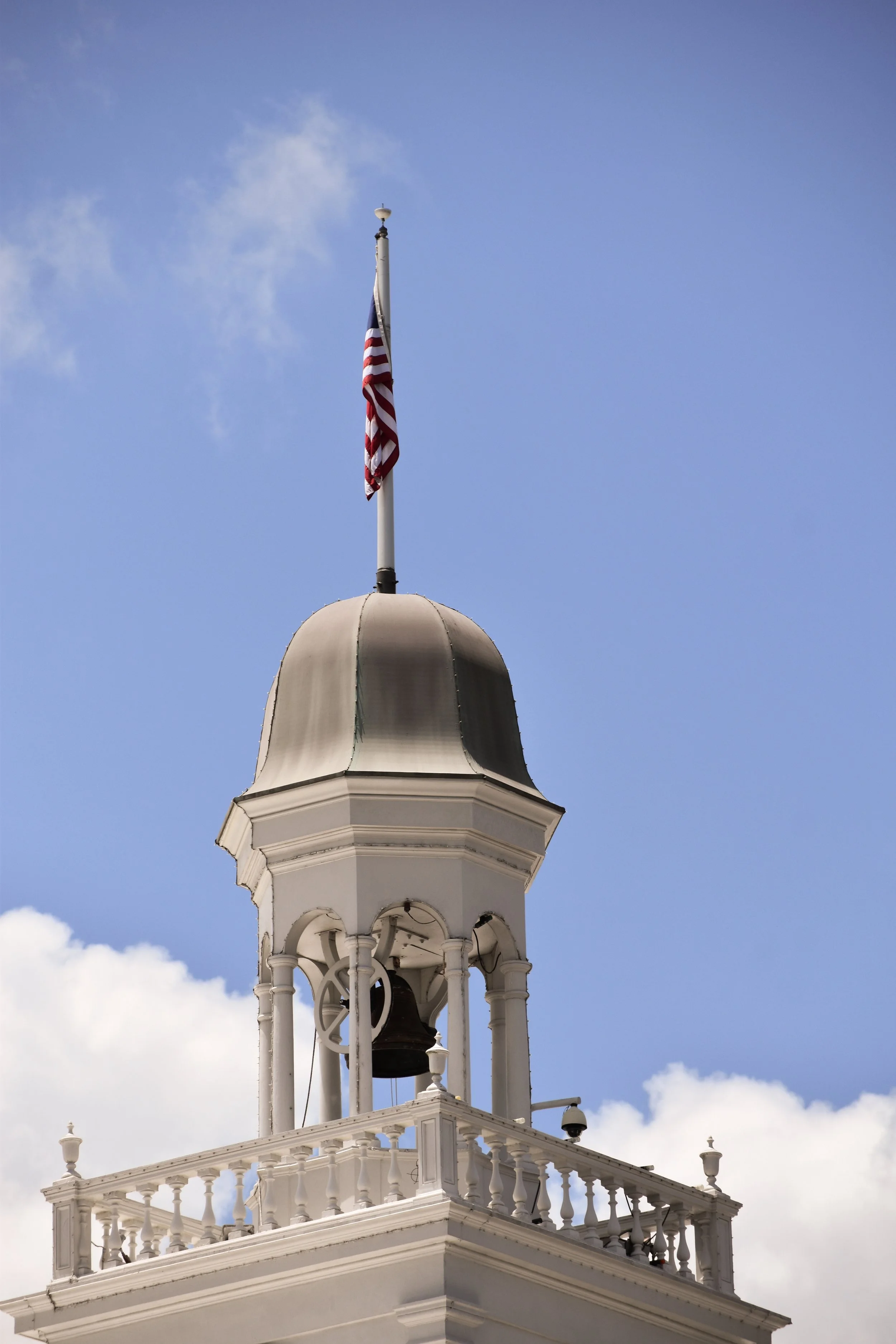 A clock tower with a bell and white decorative railing, flying an American flag against a blue sky with clouds.