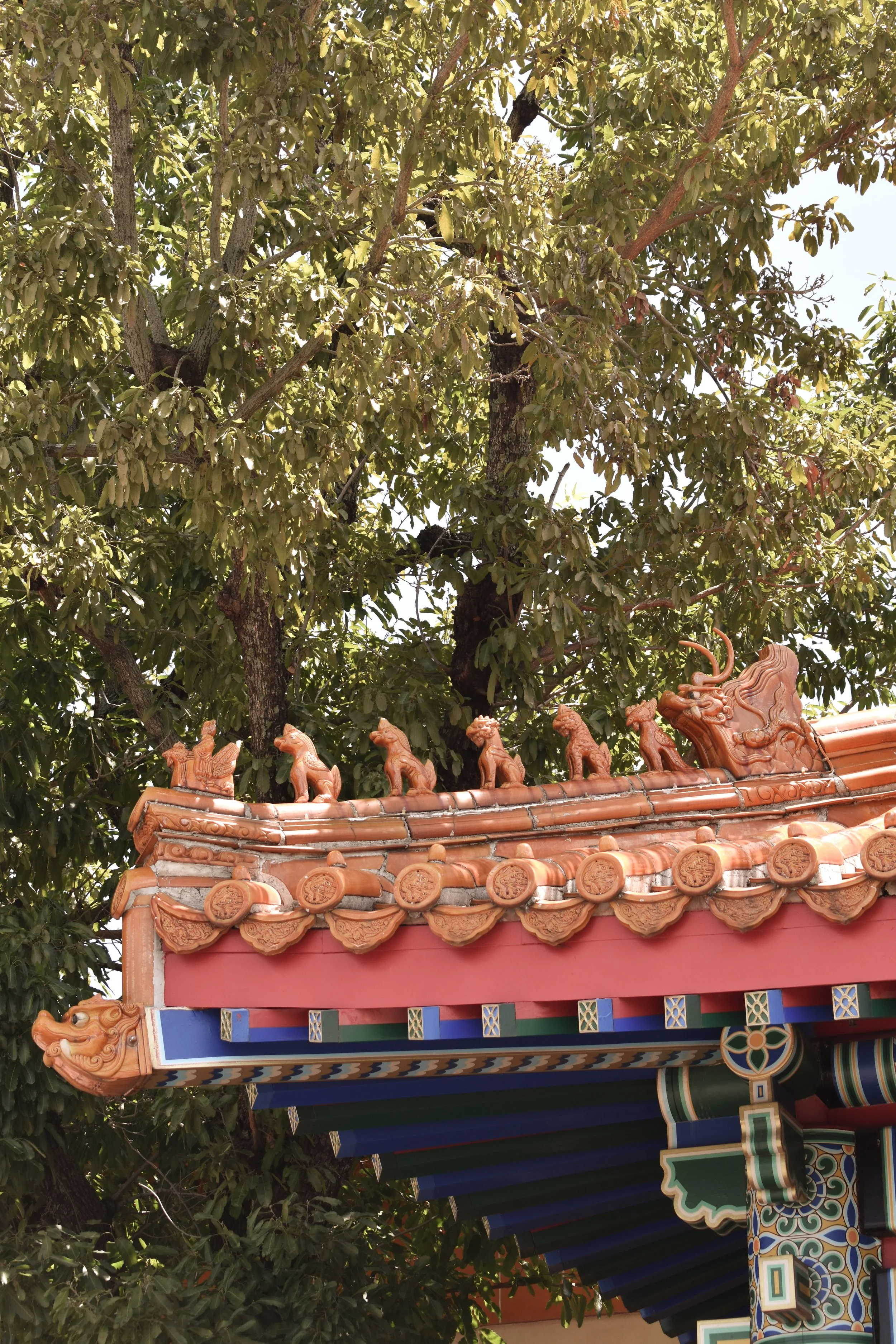 Close-up of the ornate rooftop of a traditional Chinese temple, featuring carved orange roof tiles, figurines of mythical creatures and animals, and decorative painted woodwork against a background of green foliage.