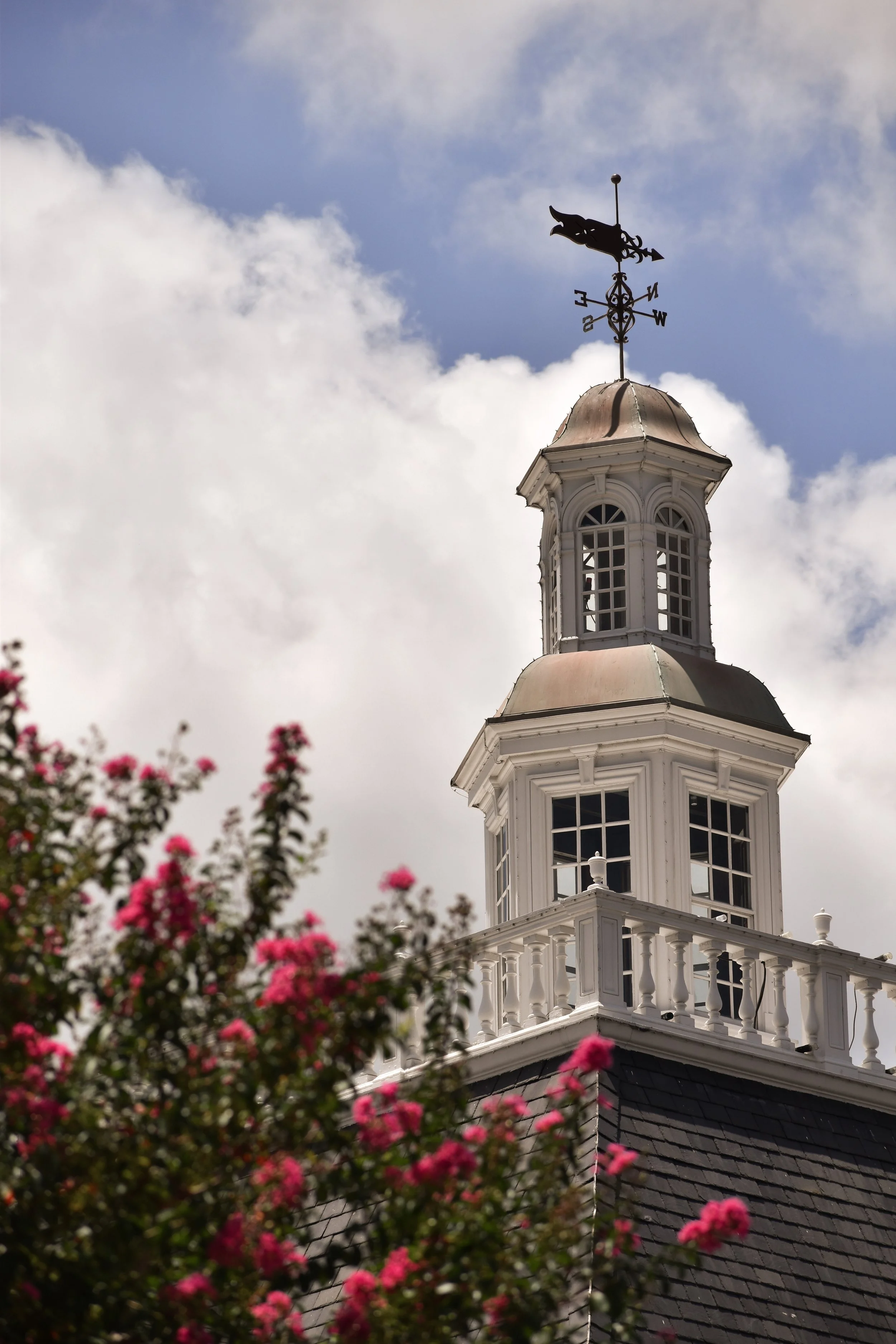 Close-up of a white church steeple with a weather vane on top, showing a rooster and directional markers, against a partly cloudy sky, with pink flowers in the foreground.