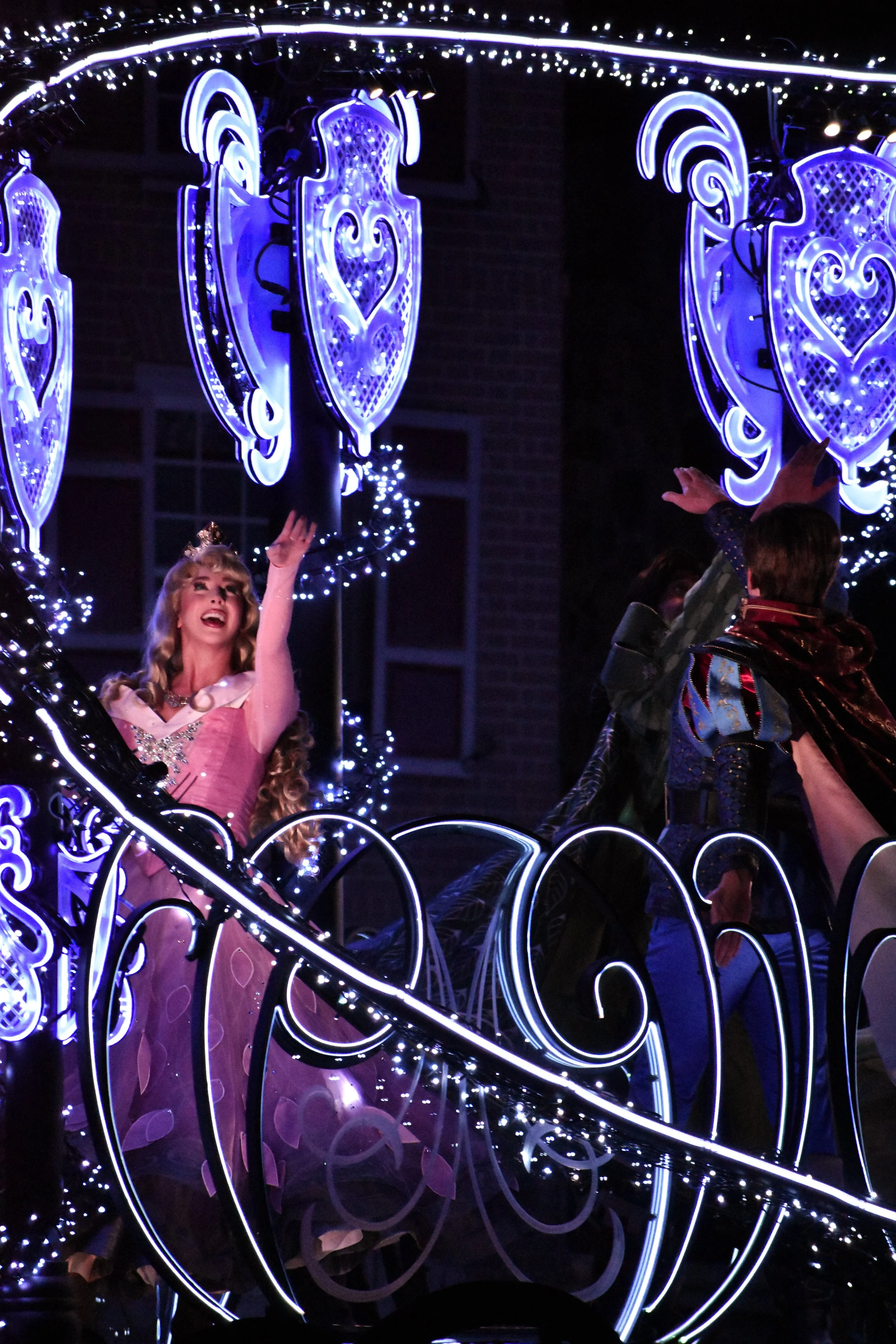 Princess in pink dress waving from a illuminated float decorated with blue and white lights.