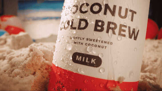 A can of coconut cold brew with milk surrounded by sand and beach.