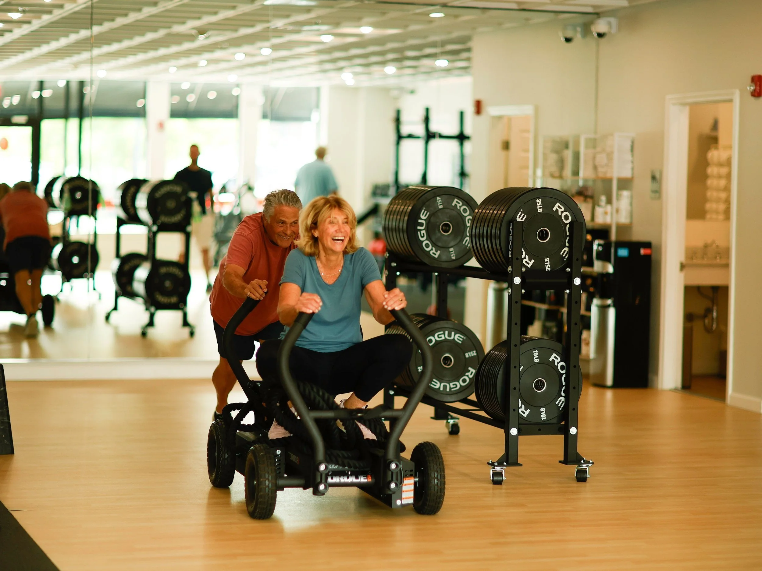 2 seniors having fun pushing and riding on a sled during strength training workout in a semi-private setting at the genesis fit