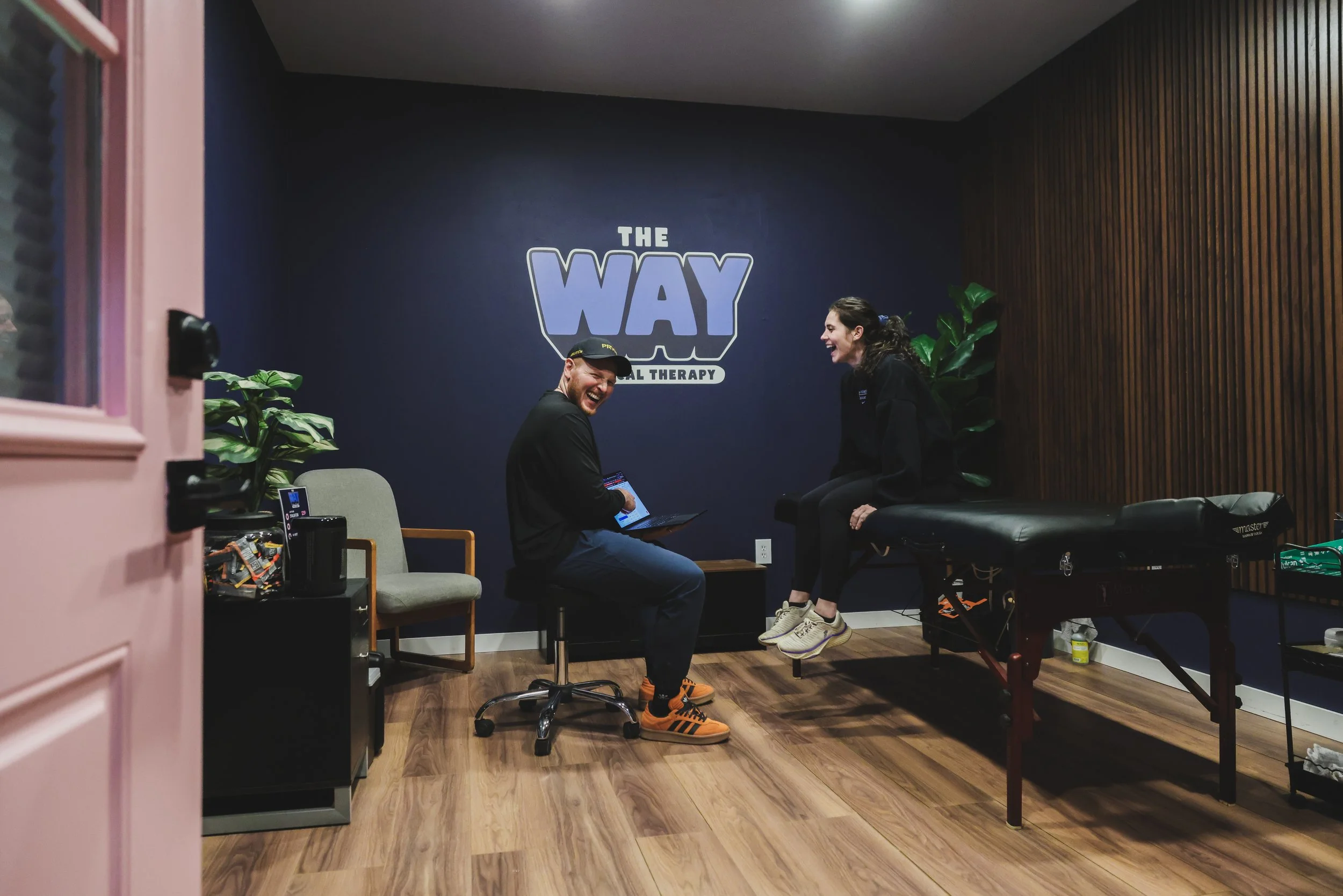 A man and woman laughing in a physical therapy clinic. The man is sitting on a chair with a laptop, and the woman is sitting on a therapy table. The clinic has a blue wall with a sign that says 'The Way Physical Therapy,' wooden flooring, and plants.