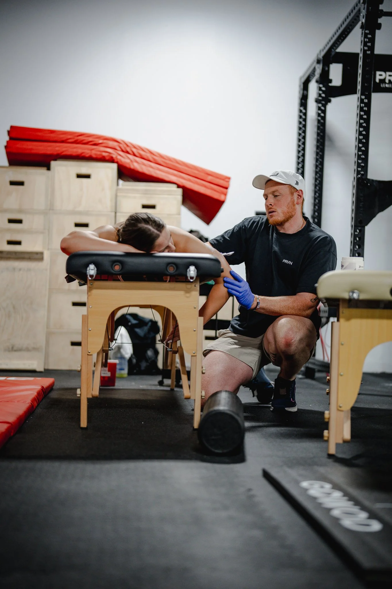 A physical therapist assists a woman lying on a treatment table in a gym, with gym equipment and mats in the background.
