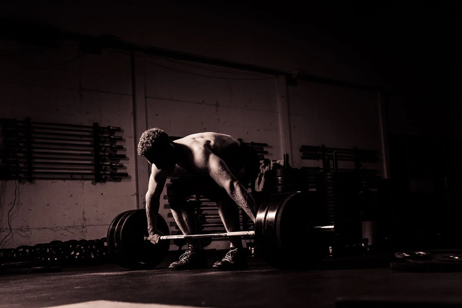 A muscular man in a gym lifting a heavy barbell with weights, with a dark background and gym equipment on the wall.