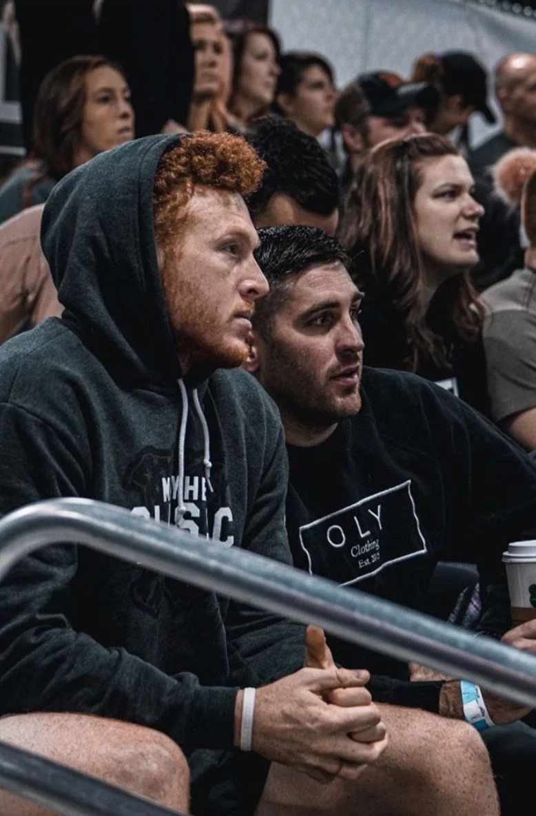 Several people seated in an audience, attentively watching an event. The focus is on two men in the foreground, one with red curly hair wearing a hoodie and the other with dark hair wearing a black hoodie, with diverse individuals visible around them.