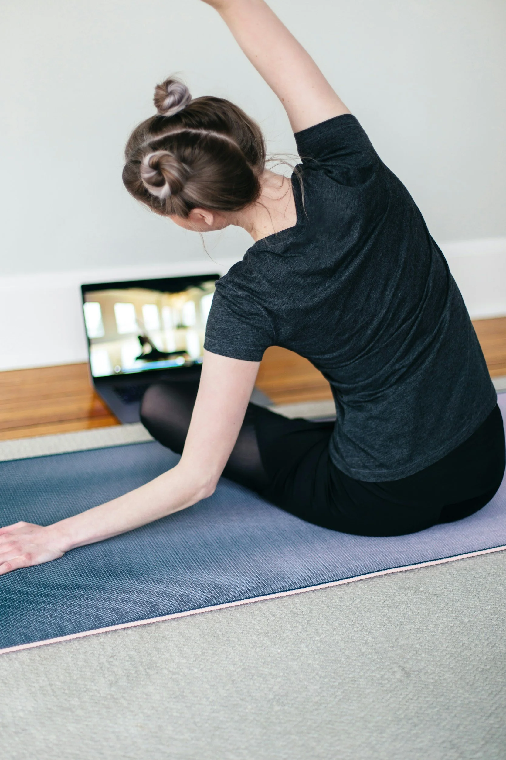 A woman practicing yoga indoors, seated on a mat in a stretching pose while watching a workout video on a laptop.