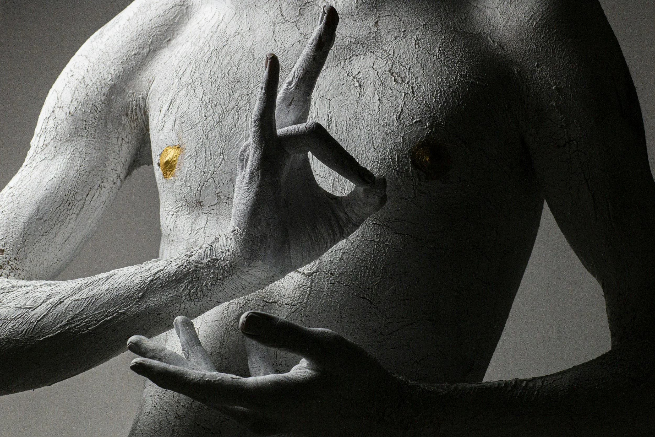Close-up of a textured, cracked gray sculpture of a human torso with one hand making an Ok gesture and the other resting below the chest area.
