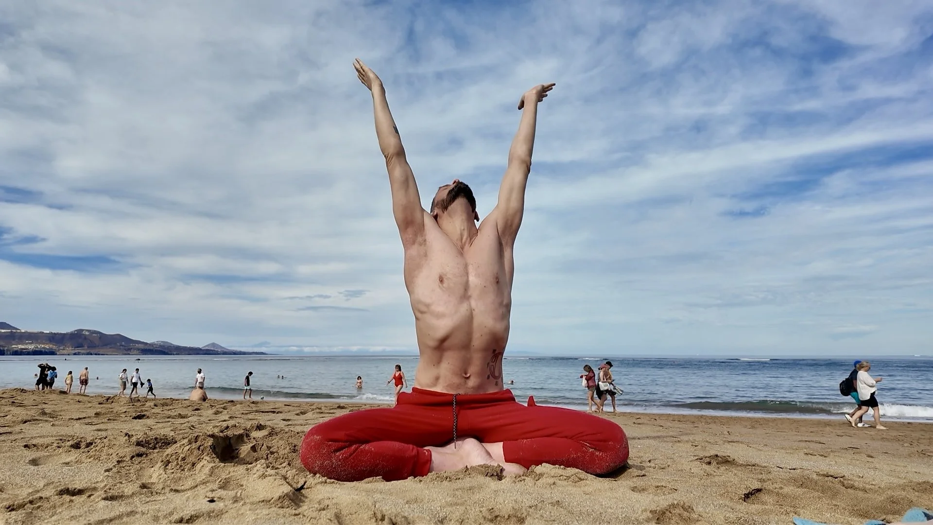 A shirtless man with red pants sitting cross-legged on the beach, practicing yoga with arms raised towards the sky, surrounded by other beachgoers near the ocean on a partly cloudy day.
