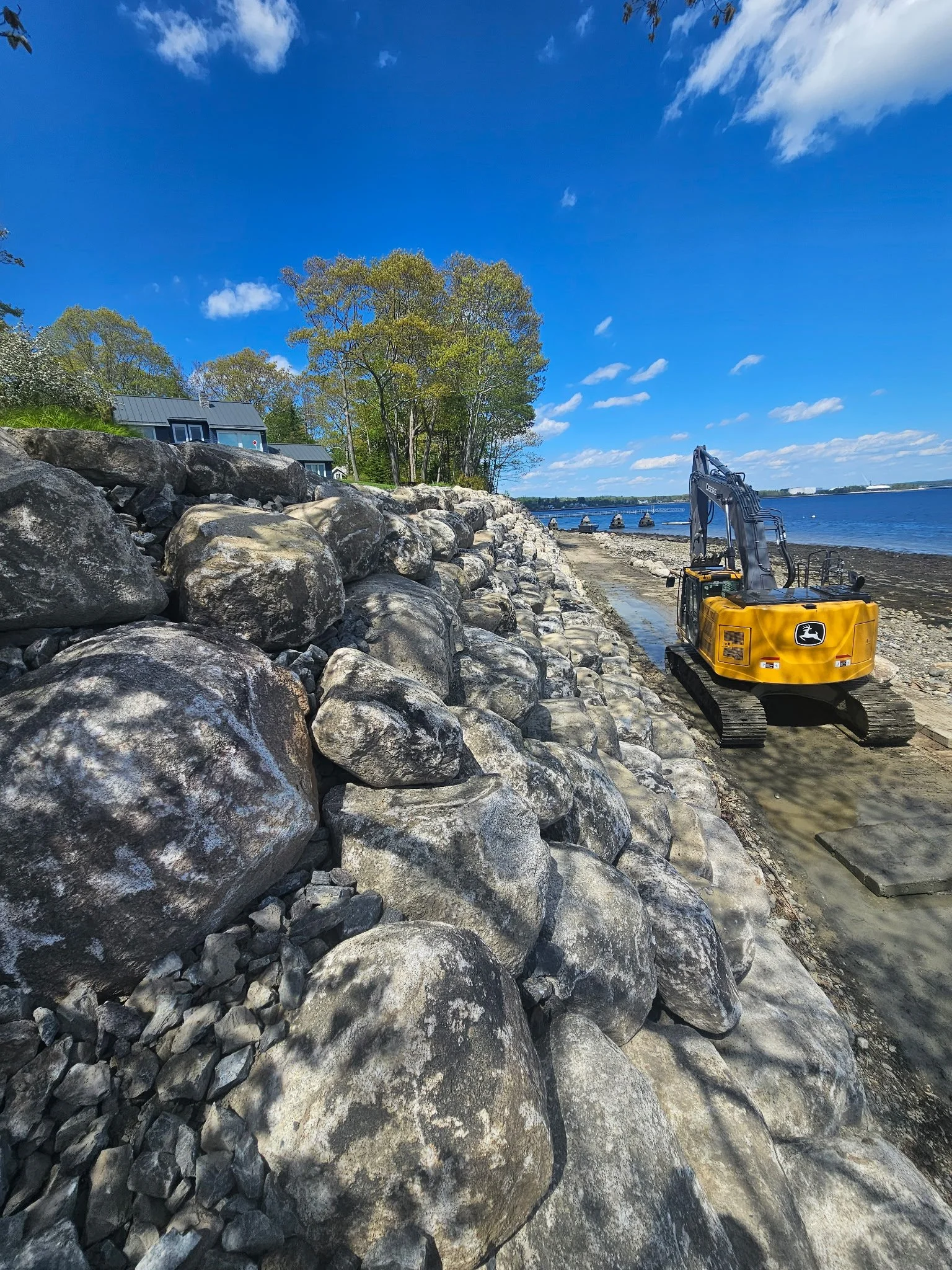 Construction equipment on rocky shoreline by a body of water with trees and a house in the background under a blue sky with clouds.
