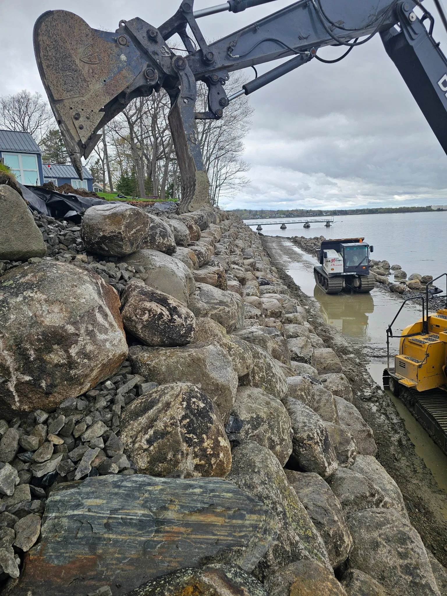 A construction site to stabilize the shoreline along the ocean with rocks and boulders being moved by new excavators. The scene features a rocky shoreline with large stones and gravel, water in the background, and cloudy skies overhead.