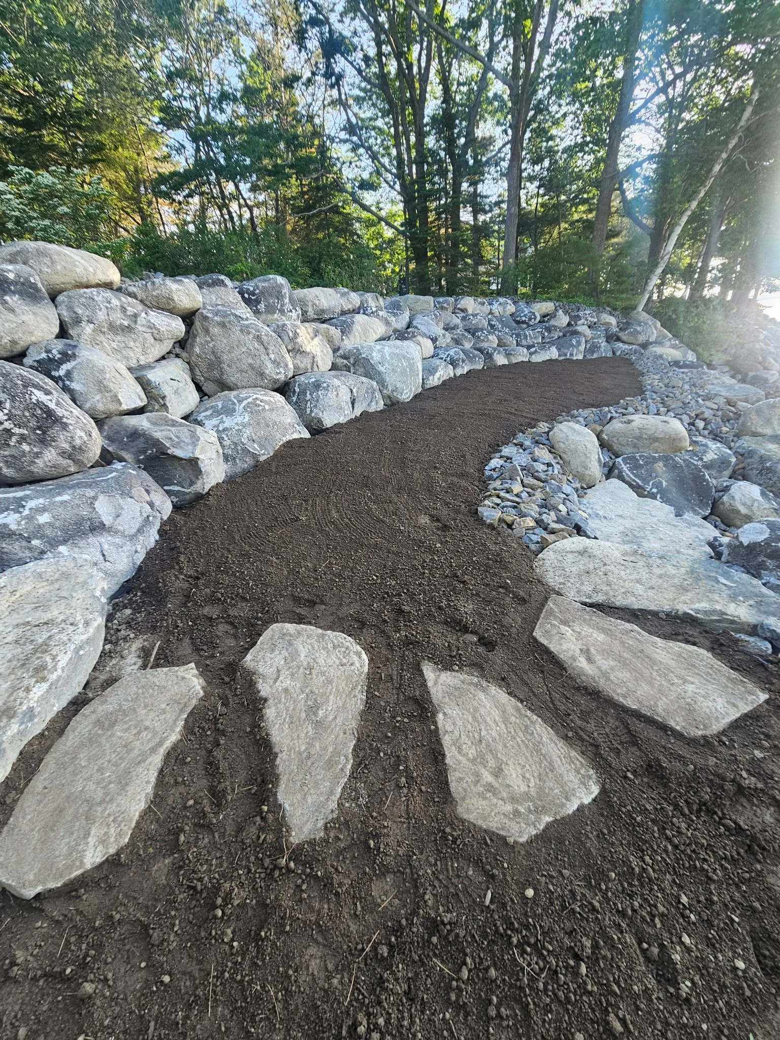 A dirt path bordered by large rocks and a stone wall, with a forest of green trees in the background.