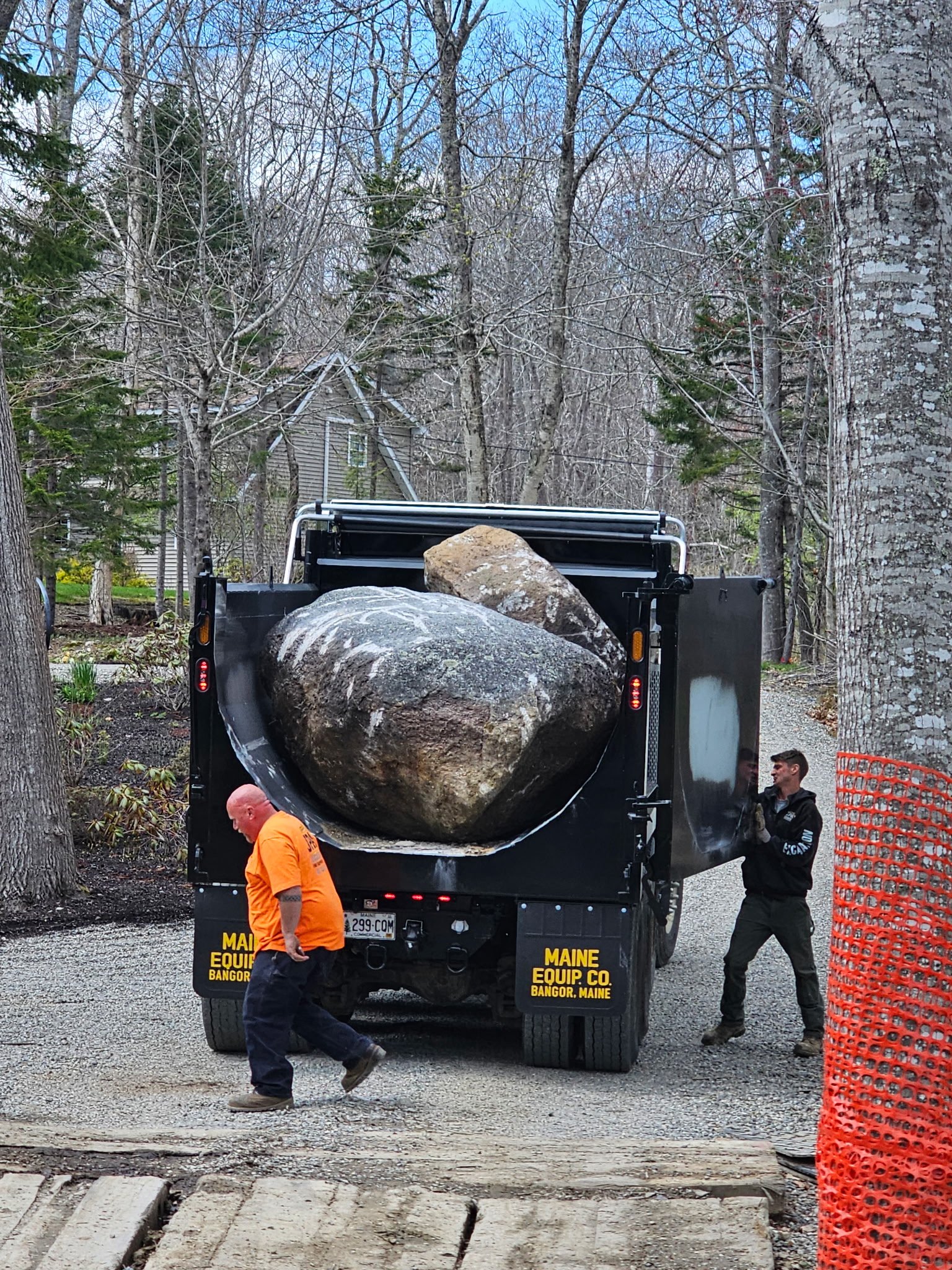 A large boulder is being transported in the back of a black truck for a seawall project. Two men are nearby: one wearing an orange shirt and the other wearing a black jacket, both walking outside among trees and construction equipment.