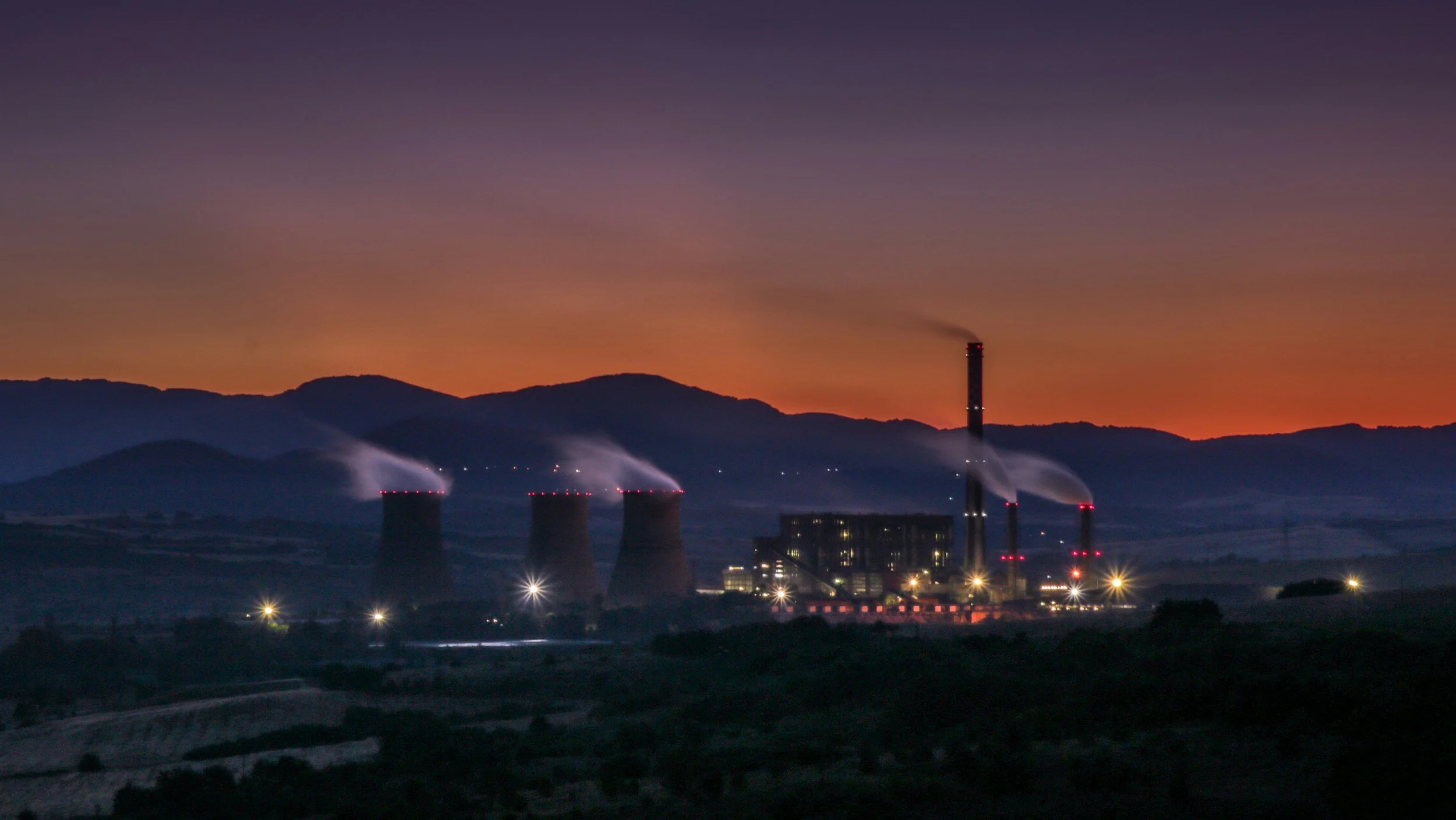 An industrial power plant with cooling towers and smokestacks at dusk, producing steam and emissions against a darkening sky.