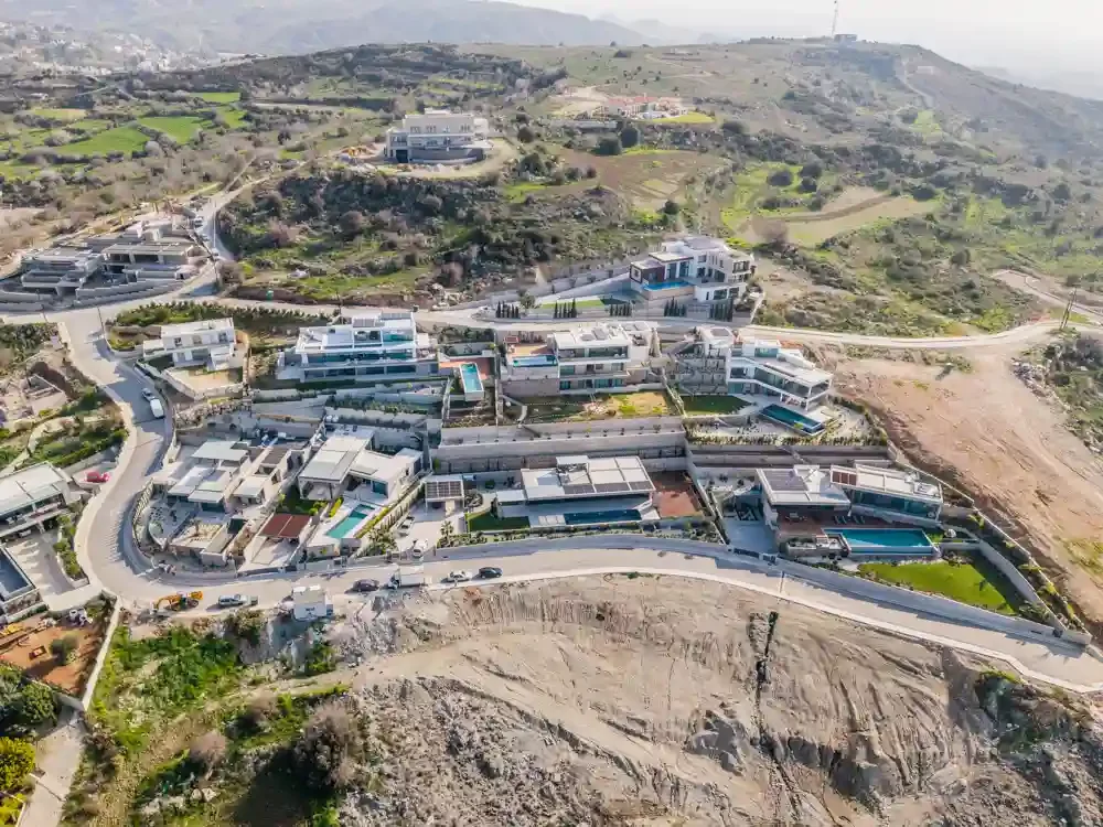 Aerial view of a hillside residential area with modern houses, some with swimming pools, and winding roads in a mountainous landscape.